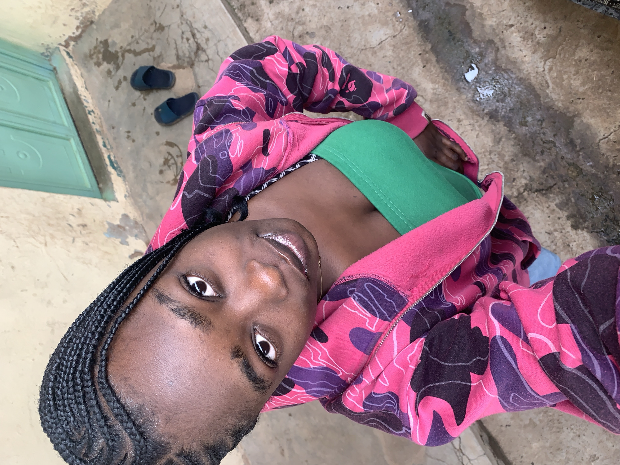 In Rafin Par, Nigeria, a young woman with braided hair and a pink and purple camouflage zip-up hoodie worn open over a green tube top is looking up at the camera. Her lips are glossed. She is standing on a dirt and concrete ground in front of a light yellow wall with a teal door. A pair of dark blue slides are on the ground near the door. Water and white residue are visible on the ground.