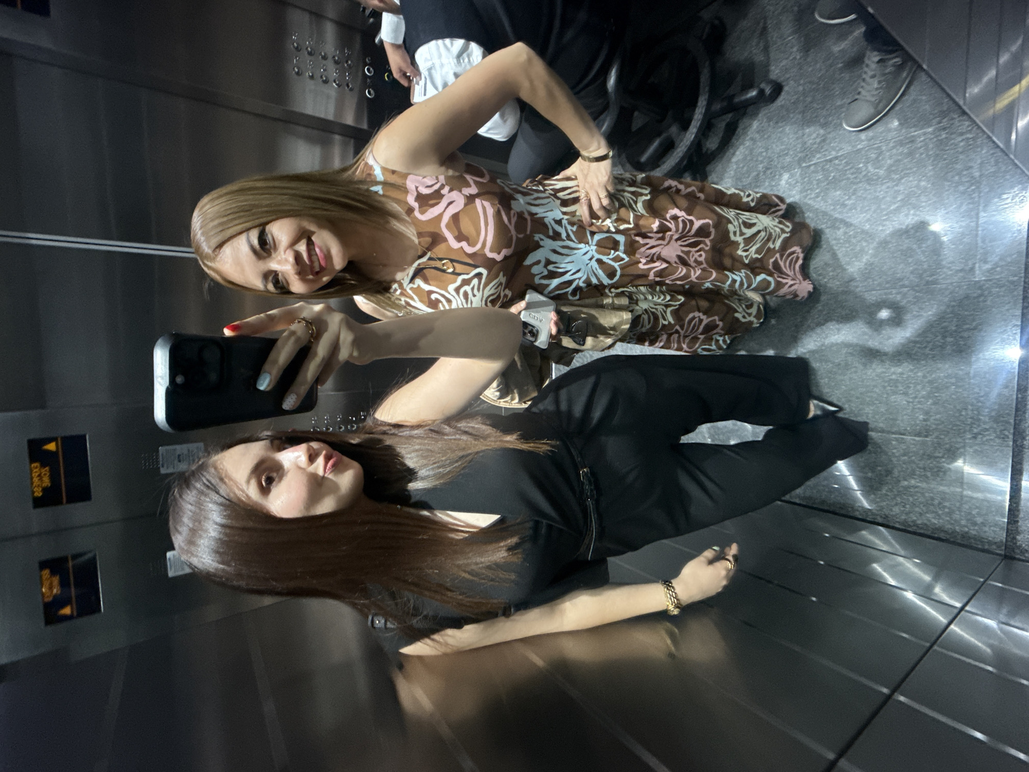 Two women are taking a selfie in an elevator. One woman is wearing a brown floral dress and the other woman is wearing a black jumpsuit. The elevator has a modern design with stainless steel walls and a tiled floor. Behind them is a person in a wheelchair. The location is potentially Mexico City, Mexico.