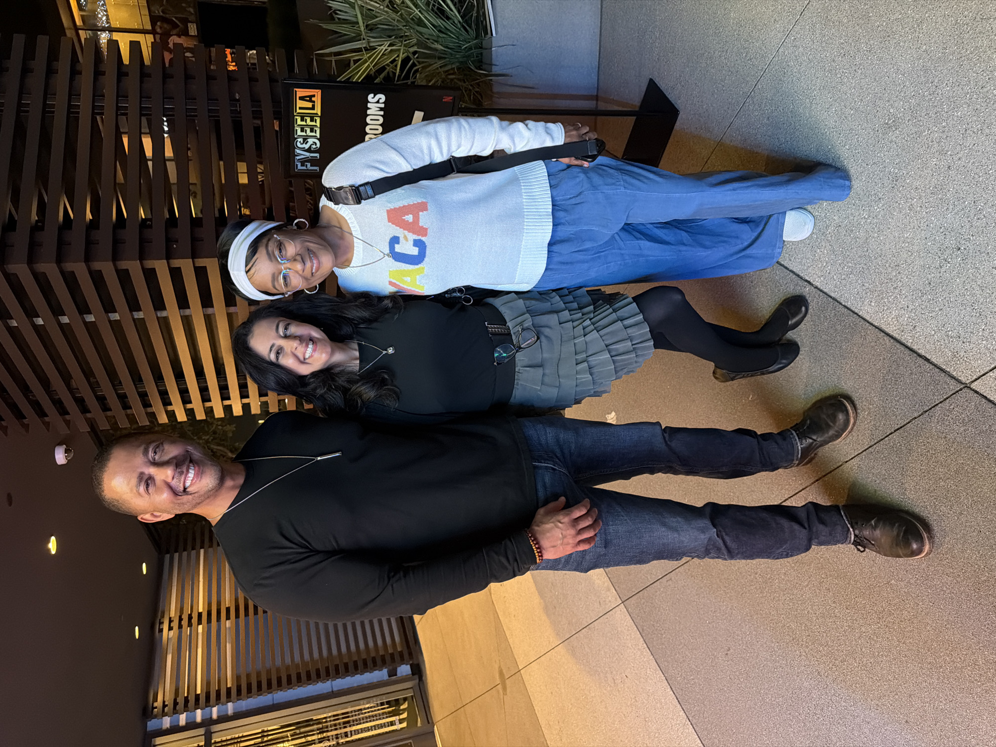 The image shows three people posing outdoors near a building at night. A man in a black long-sleeved shirt and jeans stands at the bottom of the frame. Above him, a woman in a black top, gray skirt, and black tights is positioned. At the top is a woman wearing a white sweater with colored lettering, blue pants, and a white headband. A black strap is draped across her body. Behind them is a wooden structure and a sign that reads "FYSEE LA" along with the word "ROOMS". The flooring appears to be stone or tile.