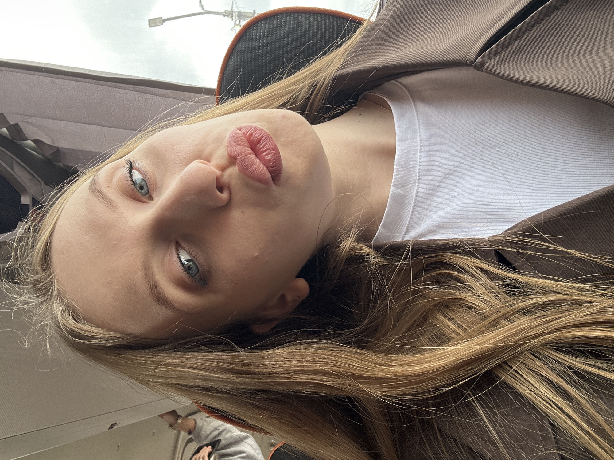 A young woman with blonde hair and blue eyes is lying down, possibly in a vehicle or train, as evidenced by the interior elements. She is wearing a white shirt and a brown blazer. Her lips are puckered, as if attempting a "duck face." The image is taken from a slightly elevated angle, looking down at her face. A partial view of another person wearing a grey sweater and an orange ring is visible in the bottom left corner of the frame. A light fixture or utility pole is visible through the vehicle window. The image does not contain enough information to confirm the location in the city of Chaikovsky, Russia.