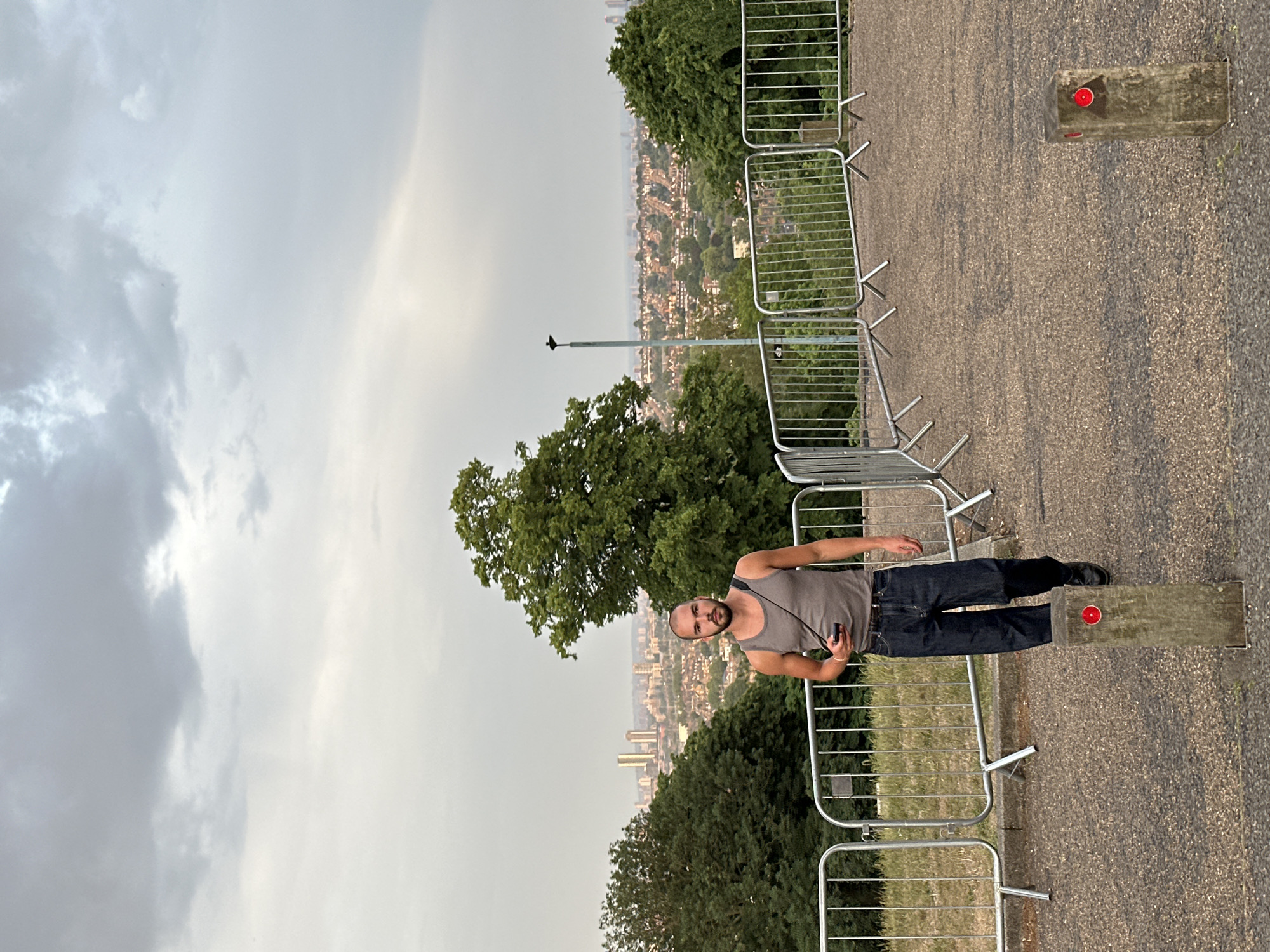 A vertical medium shot shows a man standing on a cement block along a pathway on a cloudy day. The man is light-skinned with dark hair and a beard. He wears a grey tank top, dark blue jeans, and black shoes. A thin strap crosses his shoulder. He holds an object in his left hand and looks at the camera. The pathway is lined by metal barriers. Beyond the barriers are trees and a city landscape. In the distance, tall buildings are visible. The sky is overcast with grey and white clouds. The location is Unknown, Unknown.