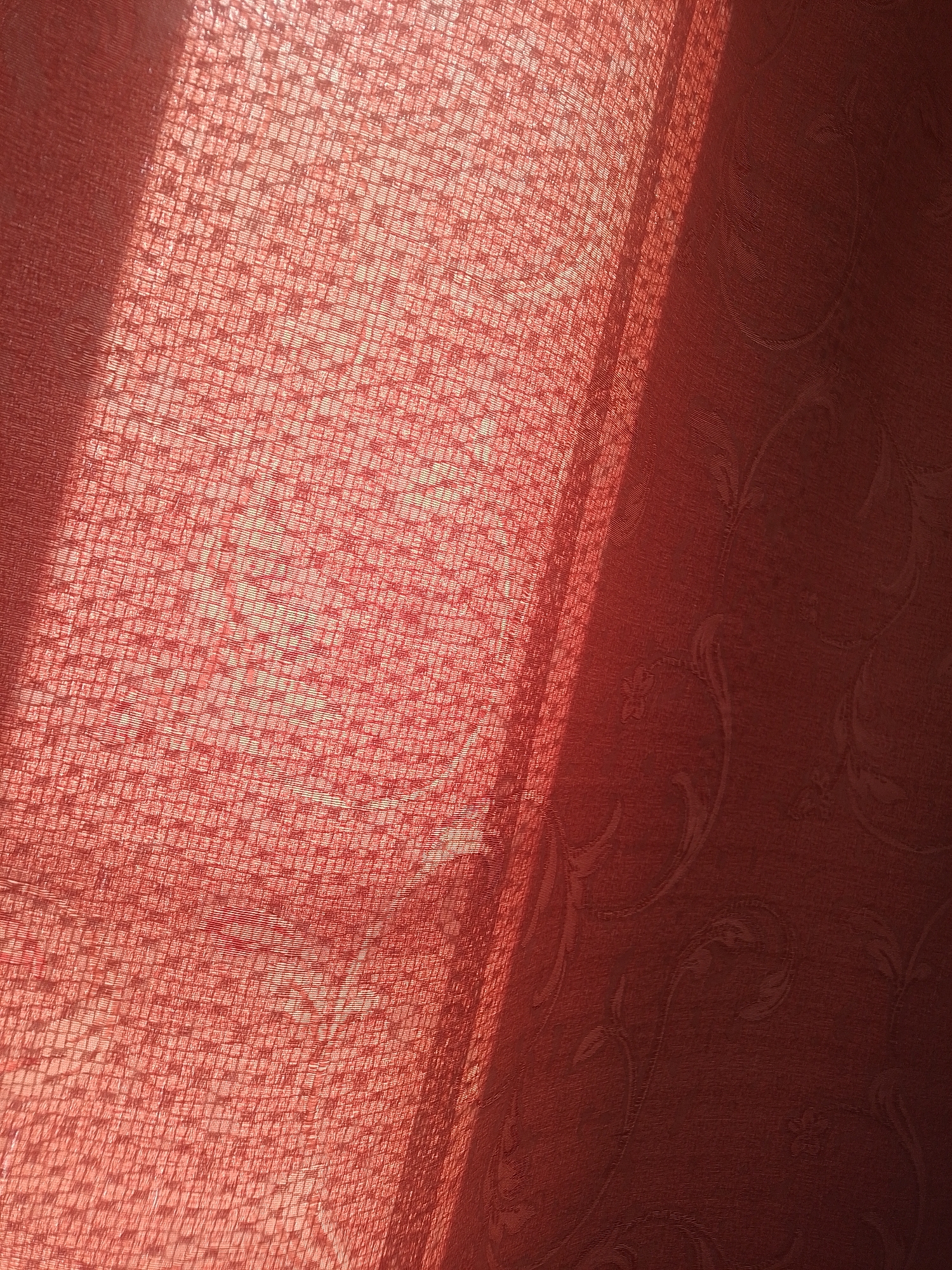 An overhead shot captures a close-up of a red textured fabric with a woven pattern. A diagonal shadow falls across the fabric from the upper left to the lower right, obscuring some of the pattern and creating a contrast between the illuminated and shadowed areas. The fabric appears to be draped, with some folds and ripples visible, particularly in the illuminated section. The texture is fine and grid-like, with individual threads clearly discernible. The color is a rich, deep red. The background is a darker shade of red, also textured, with a subtle embossed floral or paisley pattern. The lighting suggests this might be a curtain or upholstery, with sunlight filtering through or reflecting off of it. The scene is static and devoid of human presence.
