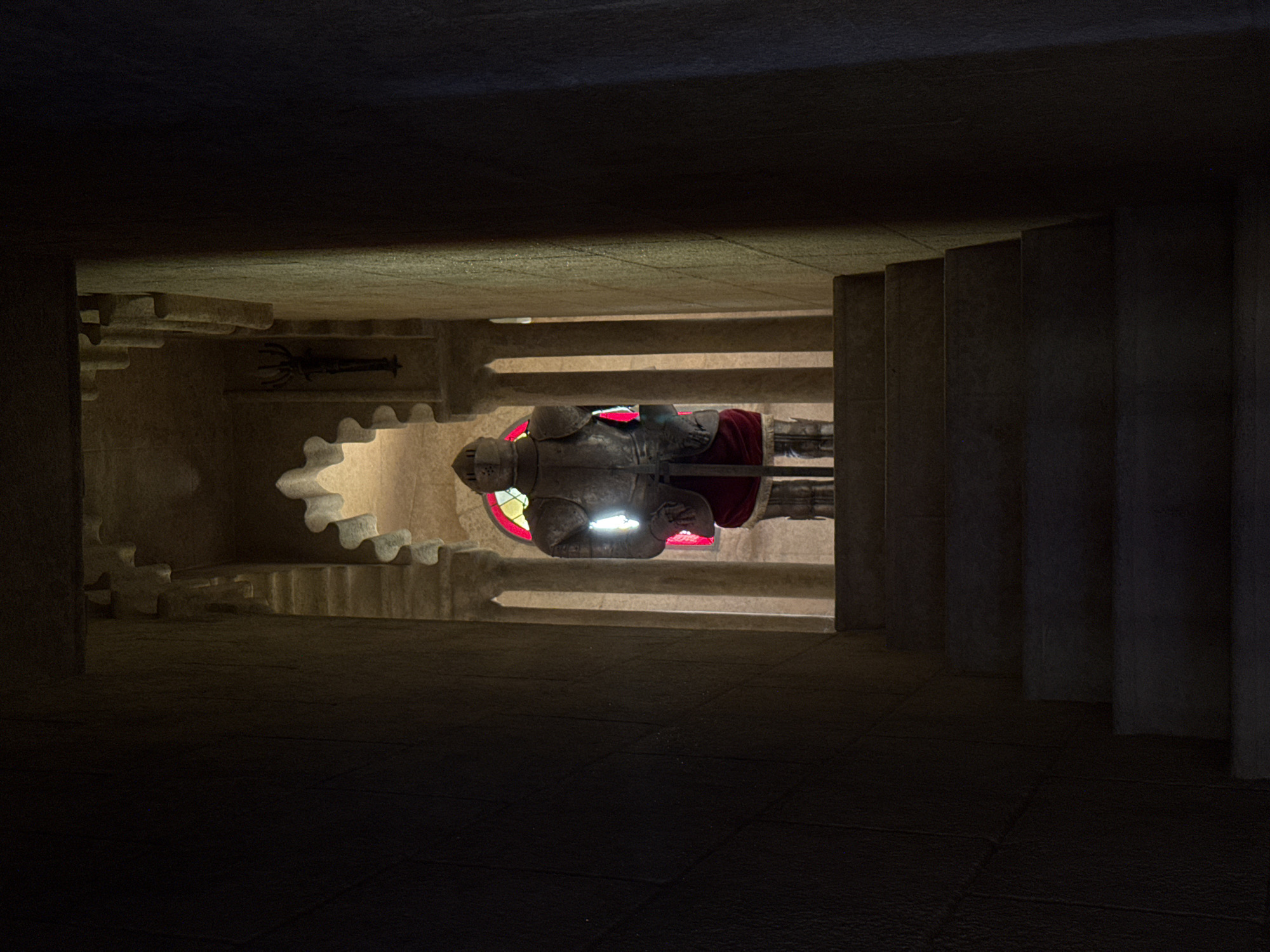 The image depicts a dimly lit, enclosed space with stone architecture. The primary subject is a statue of a knight in full armor, lying horizontally with a red cloak. The statue is illuminated by a bright light that reflects off its metallic surface. Behind the statue, a stained-glass window with red and yellow panes is visible. The setting appears to be a crypt or memorial, characterized by the rough-hewn stone walls and tiered structures that suggest stairs or seating. The overall atmosphere is somber and reverent, with the shadows and focused lighting creating a dramatic effect. The image was taken in Benalmádena, Spain, which could indicate a historical or religious significance to the site. There is no visible text in the image.