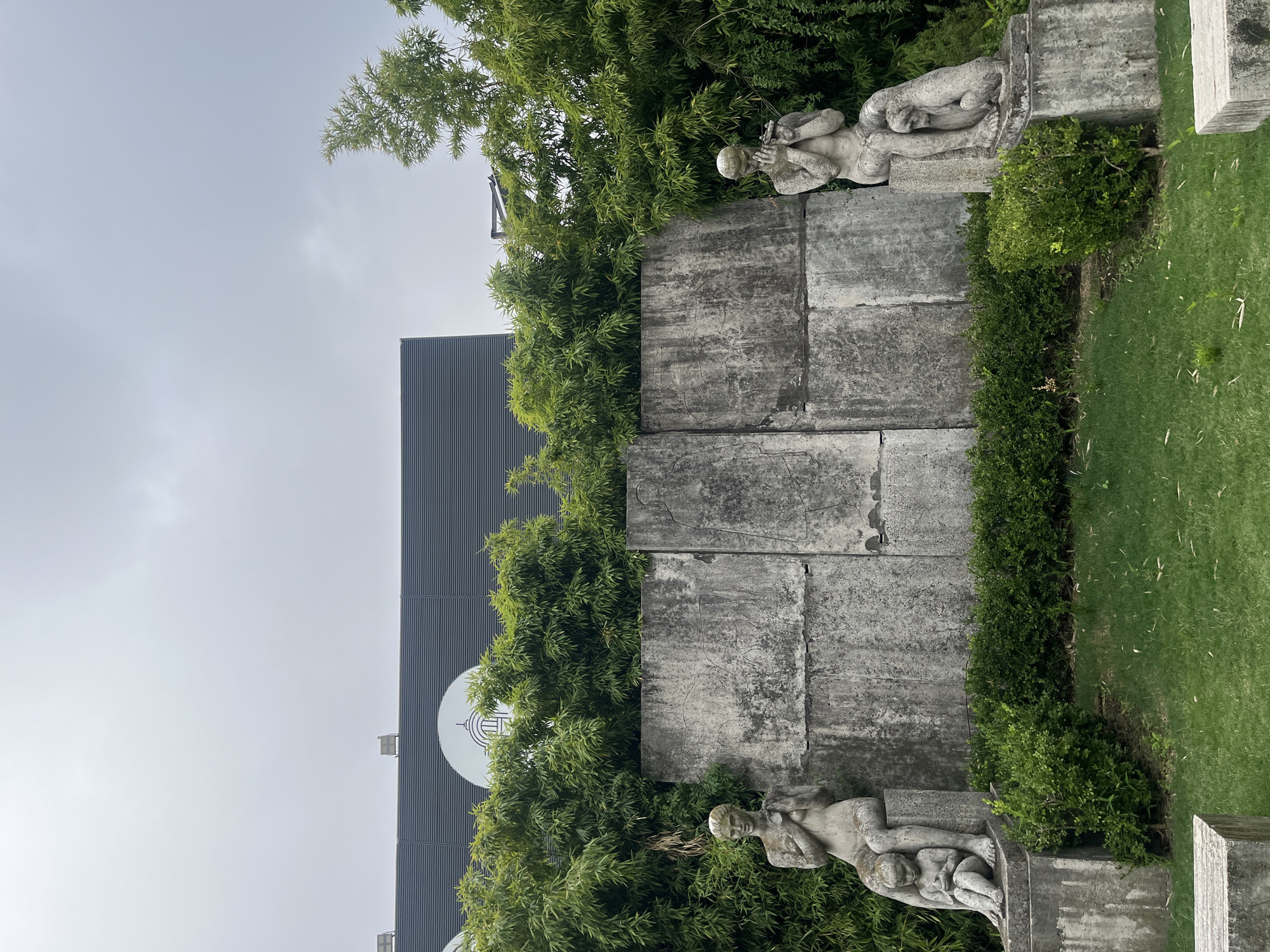 The image depicts a static outdoor scene in the Recoleta neighborhood of Buenos Aires, Argentina. In the foreground, a patch of short green grass with minimal debris is visible. Two grey concrete blocks are partially present at the bottom left and right edges. The midground features a low, manicured green shrub hedge that runs along the base of a tall, multi-paneled concrete wall, exhibiting weathering and some surface markings.
Flanking this wall are two weathered grey stone sculptural groups, each positioned on a concrete pedestal. On the left, a seated female figure is depicted with a smaller child figure seated at her feet, both facing forward. The female's hands are clasped near her chest. On the right, a seated male figure holds a flute-like instrument to his mouth, with a seated dog-like animal at his feet, both facing forward. Both larger figures appear nude.
Behind the concrete wall and statues, a dense, vibrant green barrier of bamboo foliage rises. Above this bamboo, a dark grey building with horizontal ribbed paneling is visible. A large white circular logo, depicting a stylized dome and entrance, is centrally located on the building's facade. This logo identifies the building as the Facultad de Derecho (Faculty of Law) of the University of Buenos Aires. The sky overhead is uniformly overcast and light grey. No active individuals or dynamic interactions are present.