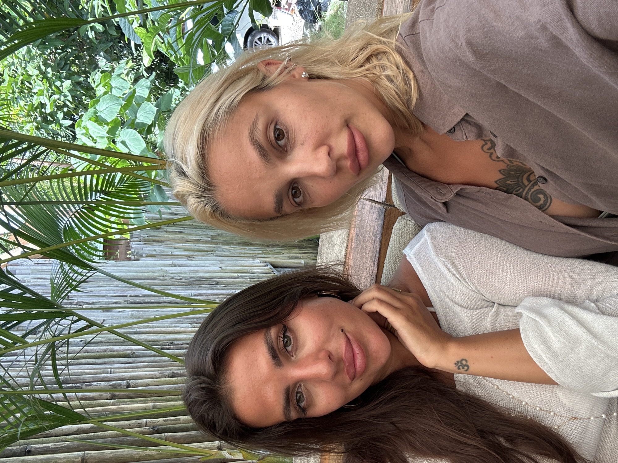 Two women are seated on a bench in an outdoor setting, with a bamboo fence and lush greenery behind them. The woman on the left has long, dark hair and is wearing a white, off-the-shoulder top adorned with a pearl necklace. She has a tattoo on her left forearm and is resting her chin on her hand. The woman on the right has blonde hair and is wearing a brown button-up shirt, revealing a tattoo on her chest. Both women are looking at the camera. The presence of palm trees and dense foliage suggests a tropical location, consistent with Kuta Selatan, Indonesia.