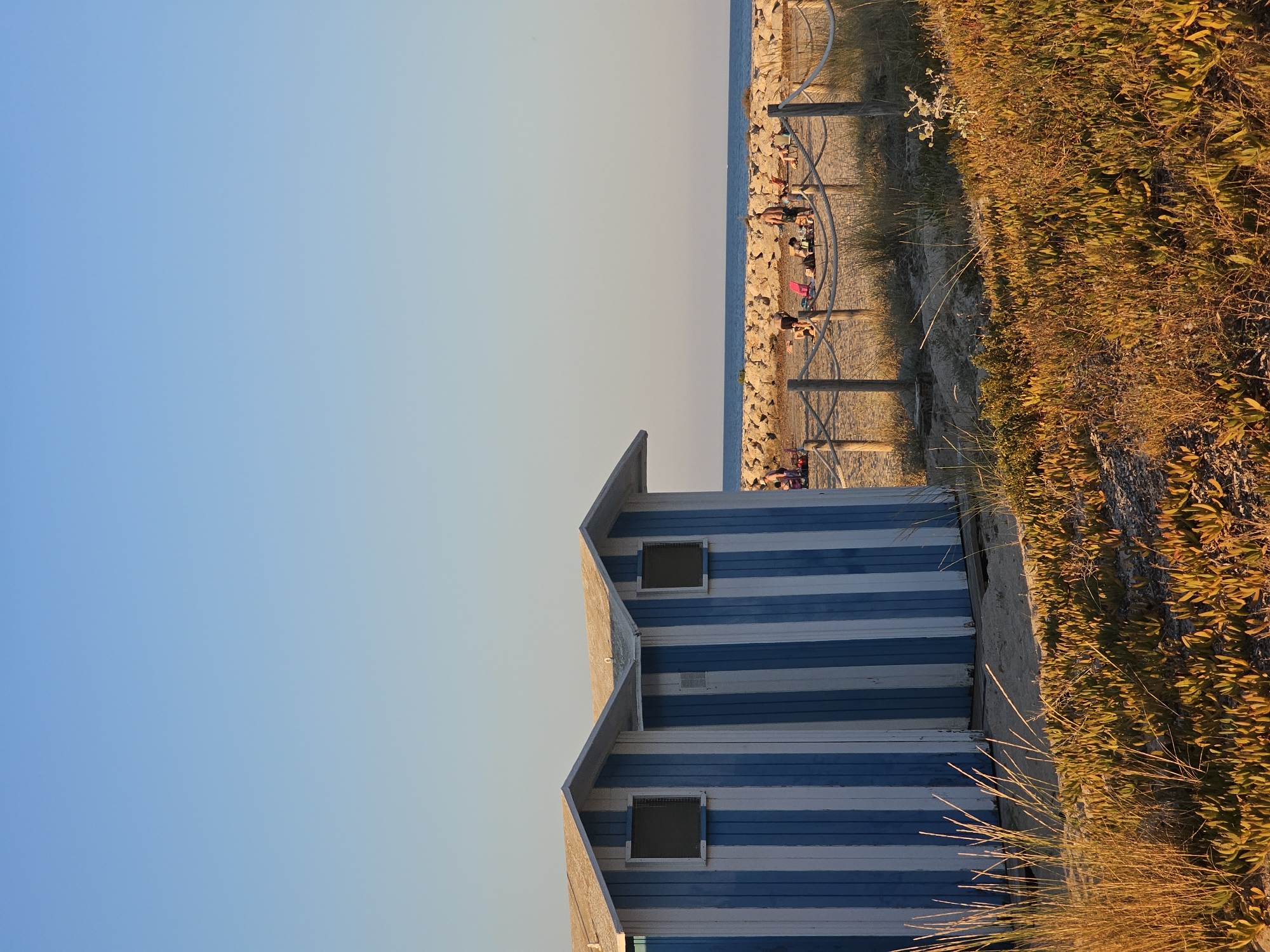 This image depicts a beach scene in Deveses, Spain, bathed in the warm glow of late afternoon or early morning sunlight. In the foreground, two beach huts with blue and white vertical stripes stand side-by-side. Each hut has a simple, pitched roof and a small, square window. Adjacent to the huts, sandy ground is covered with low-lying green and yellow vegetation, with tall, dry grass in the immediate foreground. A rope fence with wooden posts separates the huts from the beach area beyond.
In the midground, several people are scattered on the sand, some reclining on chairs and towels, others standing or walking. They appear to be engaged in typical beach activities like sunbathing or relaxing. A rough, stone breakwater runs horizontally across the scene, separating the beach from the calm, blue sea. The water stretches to the horizon under a clear, pale blue sky. The overall atmosphere is tranquil and summery.