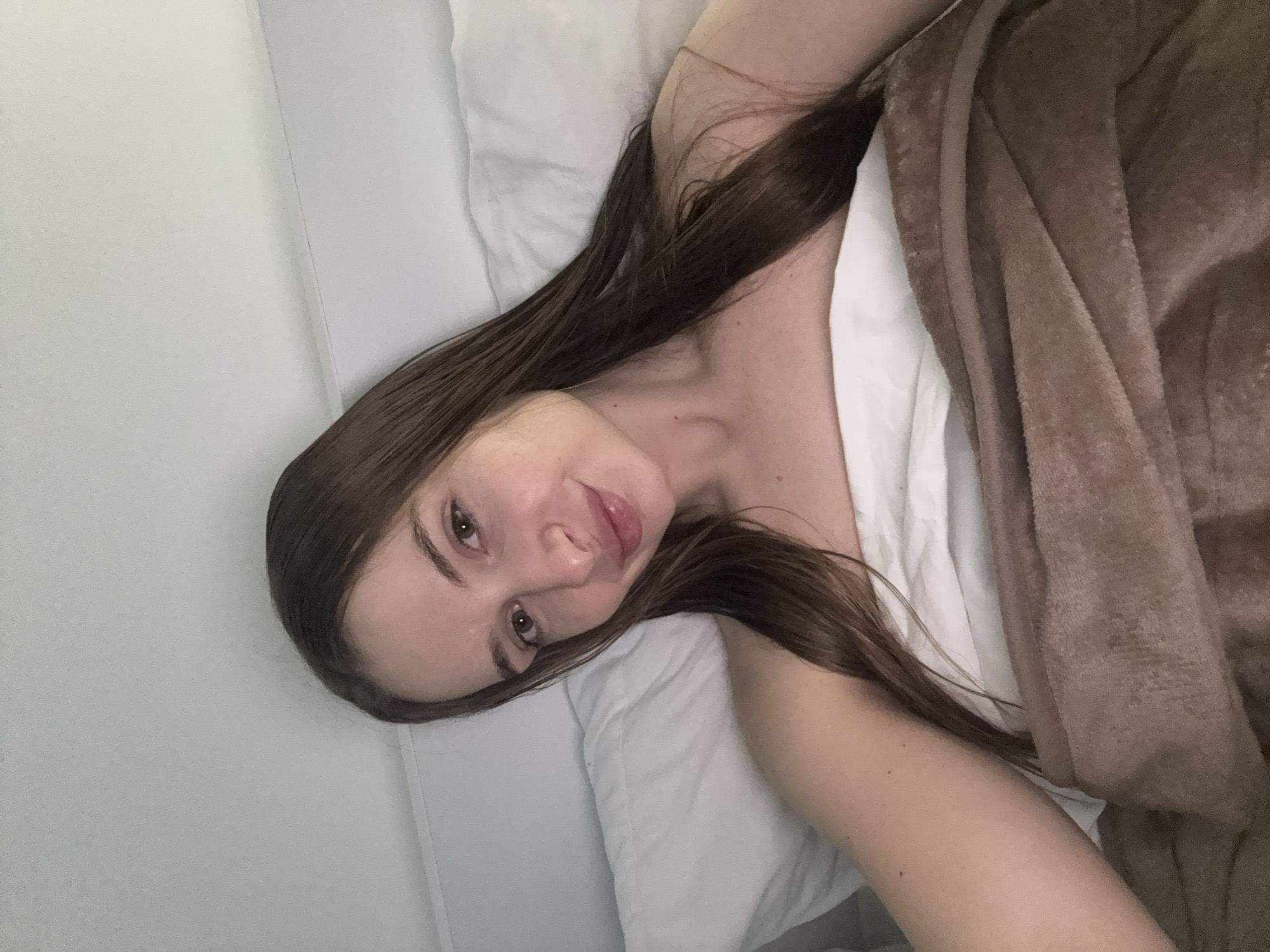 A woman lies in a bed, partially covered by a brown blanket. She has long brown hair and is facing the camera. She appears to be lying on a white pillow and white sheets. The setting is indoors. There is no specific information to discern that the location is in Budva, Montenegro.