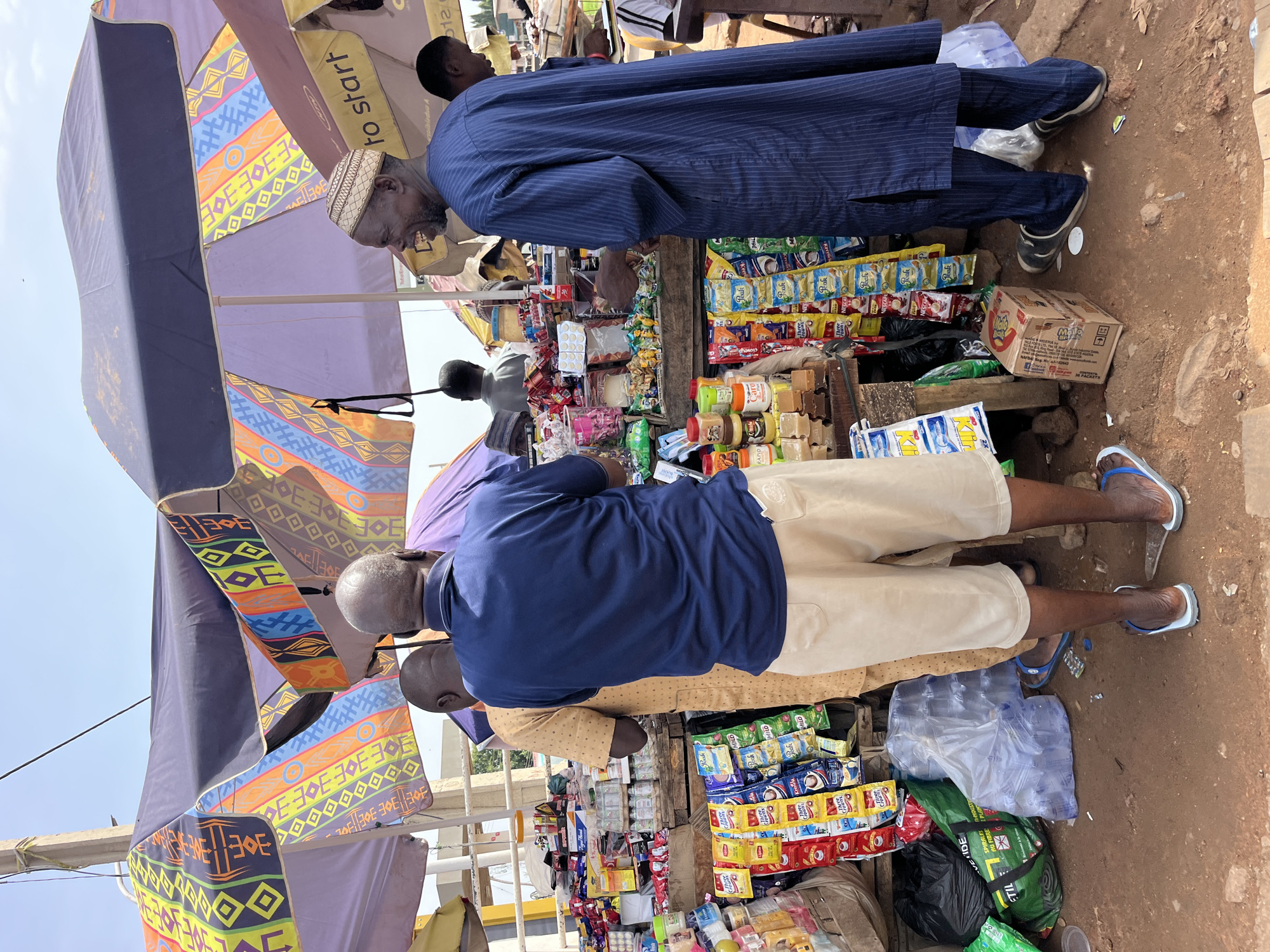 The image shows an outdoor market scene with several people. In the foreground, a man wearing a navy blue shirt and light-colored shorts stands with his back to the camera, facing a market stall. To his left, another man in a yellow garment is partially visible. To the right of the man in the navy shirt, a man wearing a blue striped garment and a cap is engaged in conversation with someone off-camera, looking to his right and smiling. Behind him, another person is visible. The market stall is filled with various goods, including packaged food items, drinks, and soaps. Several colorful umbrellas are set up to provide shade. The ground appears to be dirt. In the background, there are more market stalls and buildings, suggesting a bustling commercial area.