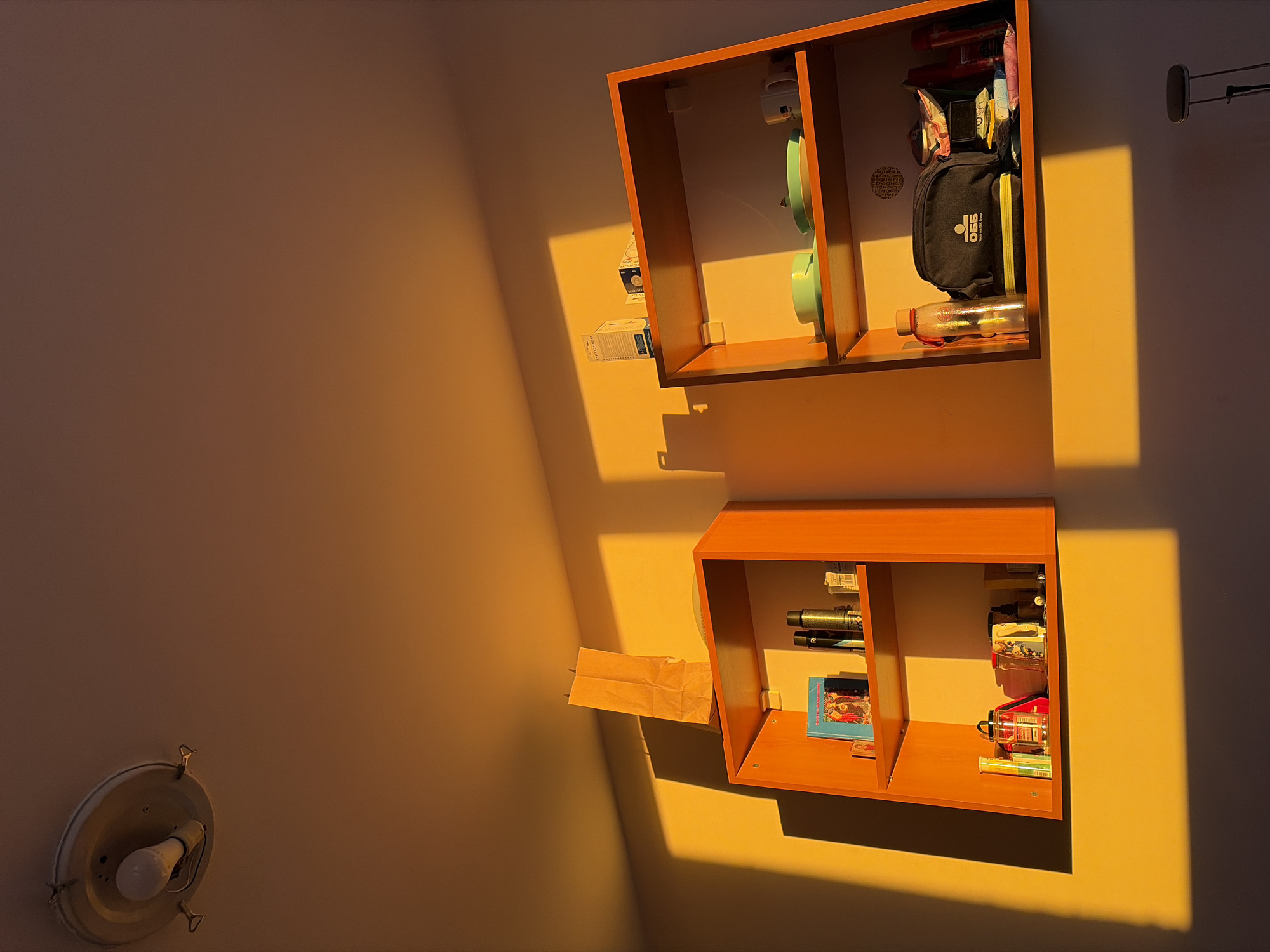 The image depicts two open-backed, wooden shelf units mounted on a wall. The upper unit contains objects including teal-colored items, a black bag with a green zipper, and a clear water bottle. The lower unit contains objects including pens, a book, and stacked tins. A brown paper bag rests on the wall below the lower shelf unit. The wall is painted a light color, and the scene is heavily lit with warm, golden sunlight that creates sharp shadows. A light fixture is visible on the lower left. The location is not discernible from the image.