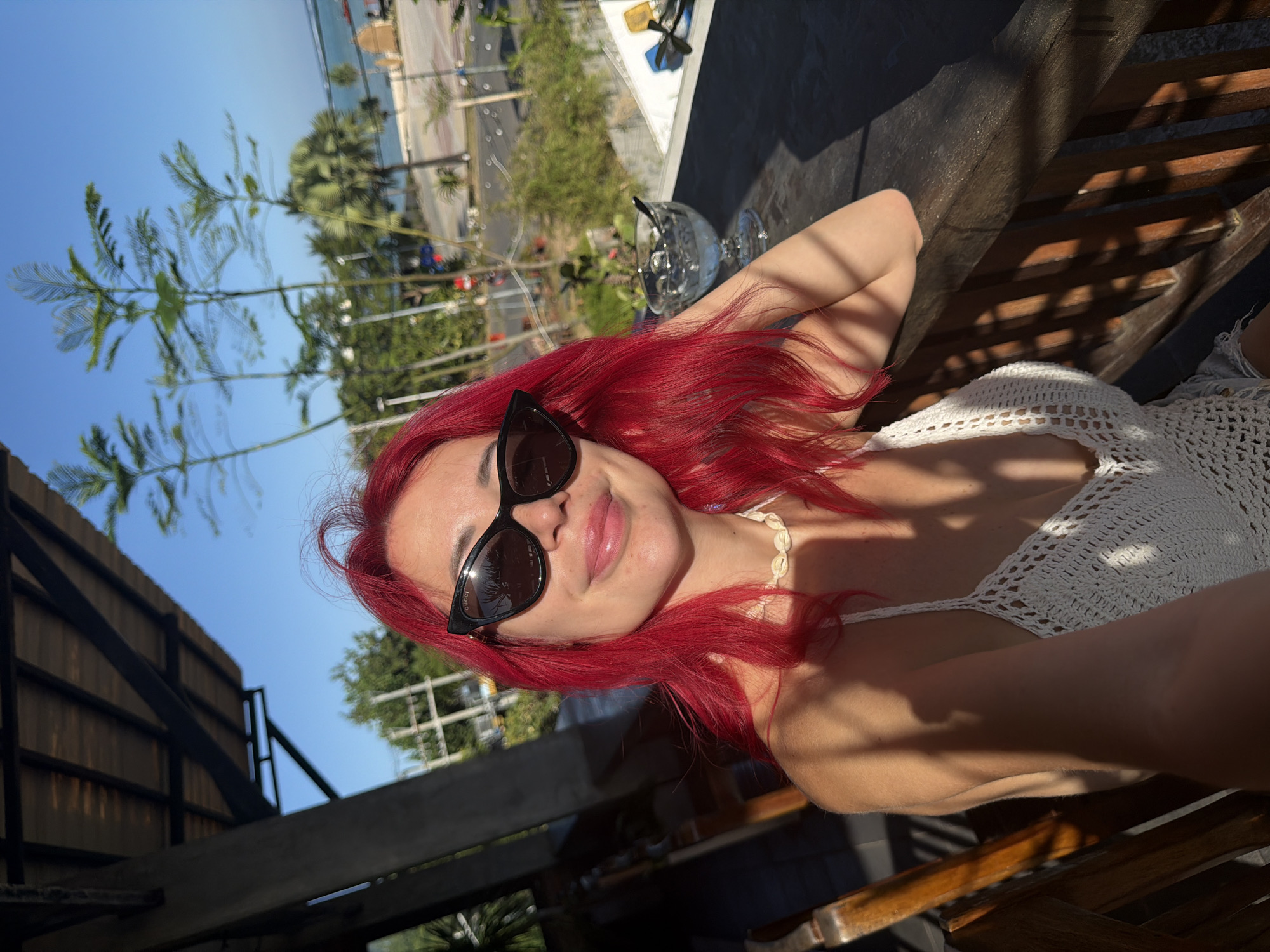 A woman with bright red hair and black cat-eye sunglasses is taking a selfie while sitting at a table outdoors. She is wearing a white crochet top and a shell necklace. In the background, there is a body of water, palm trees, and a clear blue sky. A glass of ice water with a spoon is on the table next to her. The setting appears to be a restaurant or cafe in Kuta, Indonesia, likely overlooking a street and the ocean.