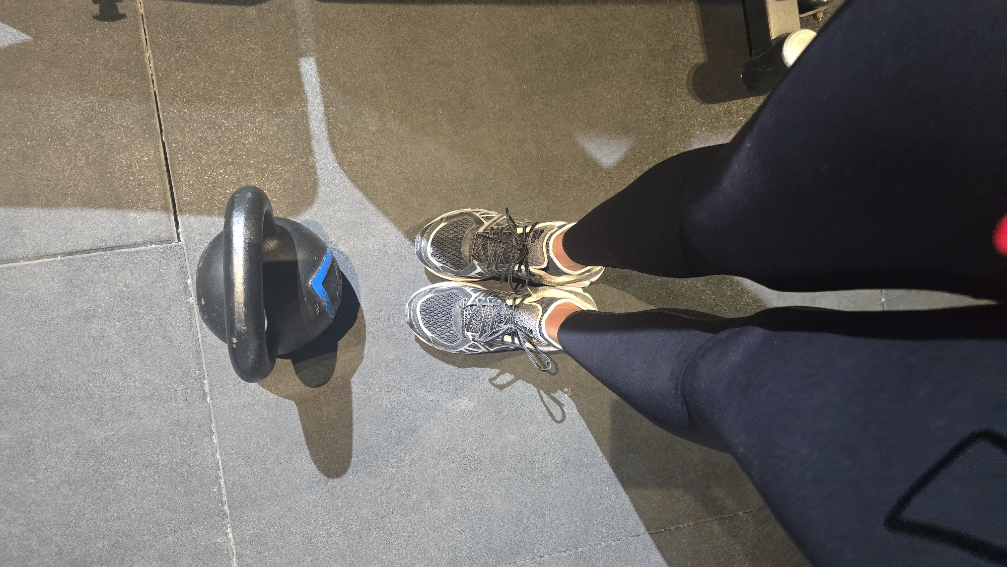 A first-person perspective image shows a person's legs and feet wearing black leggings and athletic shoes standing on a grey gym floor. A black kettlebell with a blue accent is positioned on the floor in front of the person. The scene is lit, casting shadows on the floor. The location is identified as Trikomo, Northern Cyprus / Cyprus.