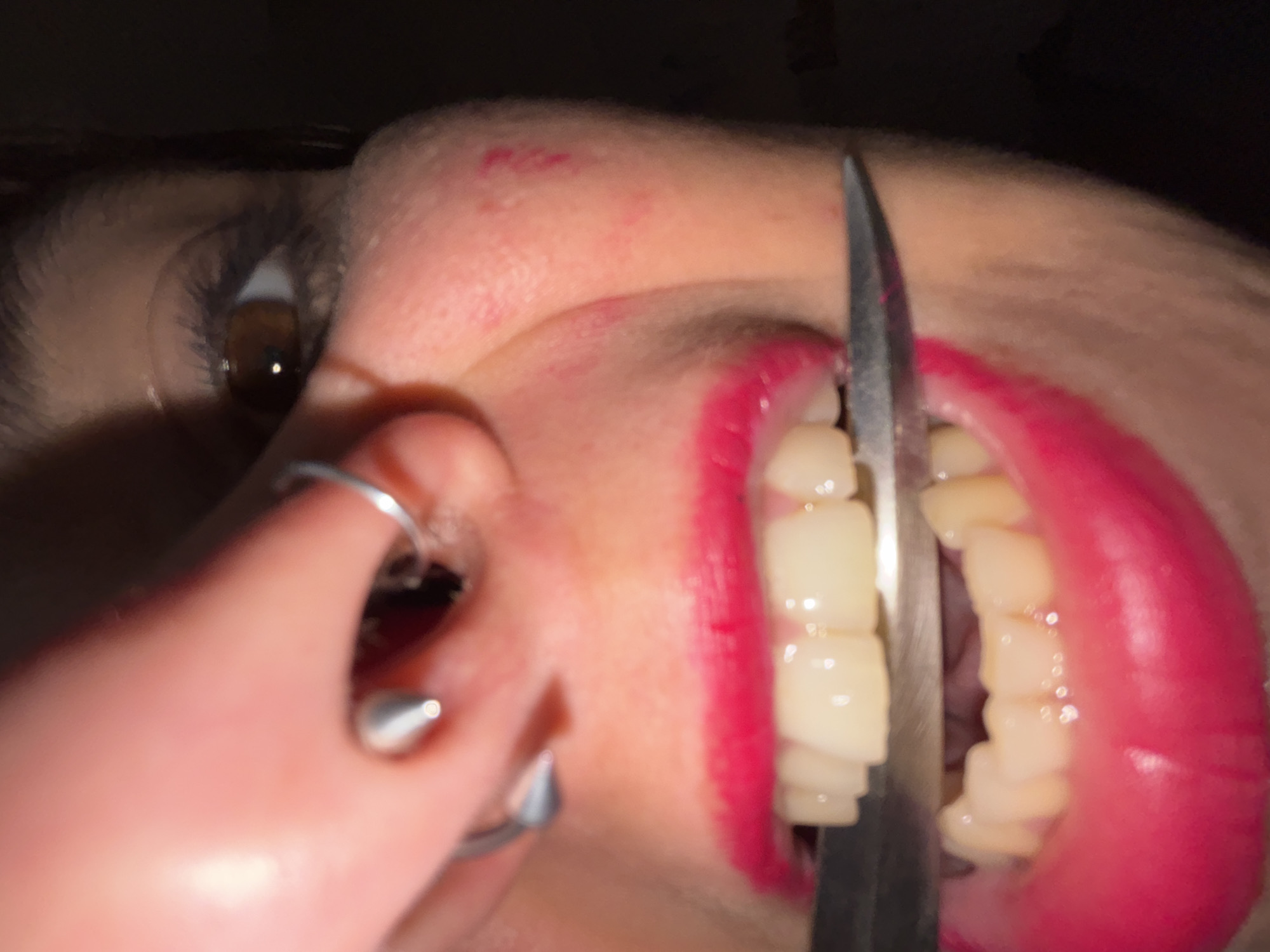 A close-up shot shows a person's face with a piercing in their nose and a knife positioned between their teeth. The person's lips are painted bright pink. The lighting is dim and the background is dark and out of focus. There is no evidence to suggest the location is Sofia, Bulgaria.