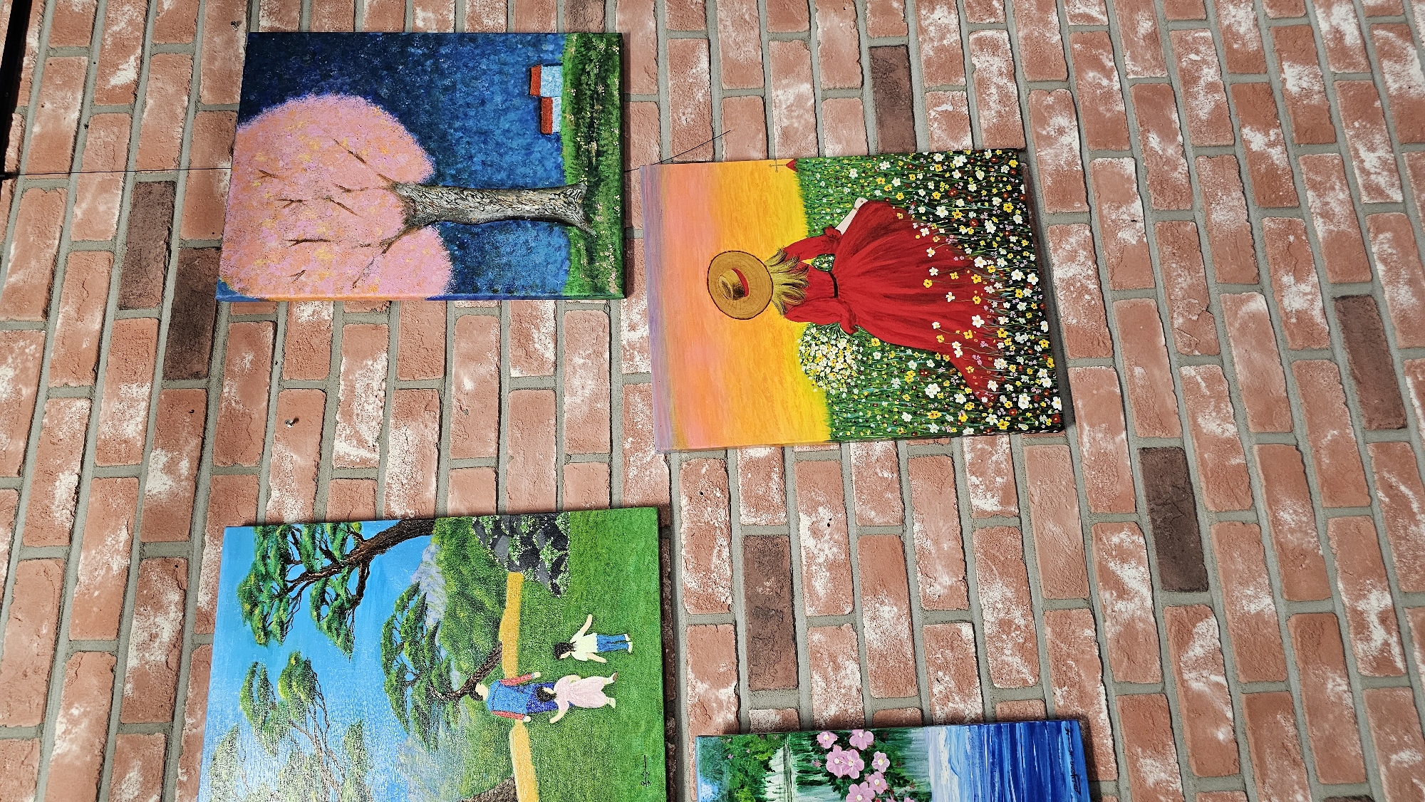 The image shows a brick wall with several paintings displayed on it. One painting on the upper left depicts a person and two children walking in a green field with trees under a blue sky. To the right of this painting is another one featuring a tree with pink blossoms against a dark blue background, with two small houses in the distance. Below the first painting and to the right of the second is a painting of a woman in a red dress with a straw hat, standing in a field of flowers. To the far left, a portion of another painting is visible, showing a body of water with pink flowers. The paintings are hung on a red brick wall, suggesting an indoor or outdoor art display, possibly in Asan-si, South Korea.