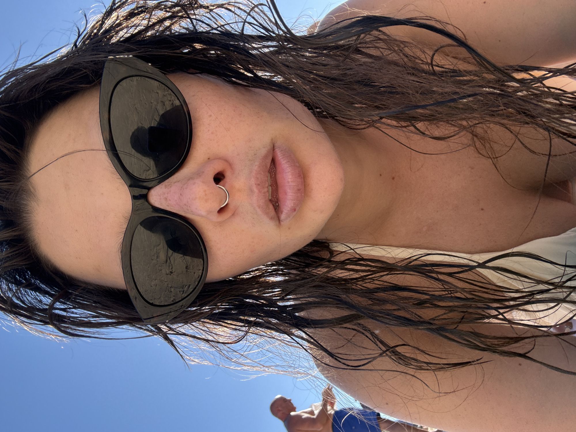 A close-up, low-angle, point-of-view shot captures a woman with wet, dark brown hair and freckles across her nose and cheeks. She wears black, cat-eye sunglasses and a light-colored bikini top. A silver septum piercing adorns her nose. Her lips are slightly parted, revealing a hint of moisture. The background shows a clear blue sky and a blurred figure of a man in blue swim trunks in the distance, suggesting a beach setting, possibly in Tel Aviv, Israel. The overall lighting suggests a sunny day.