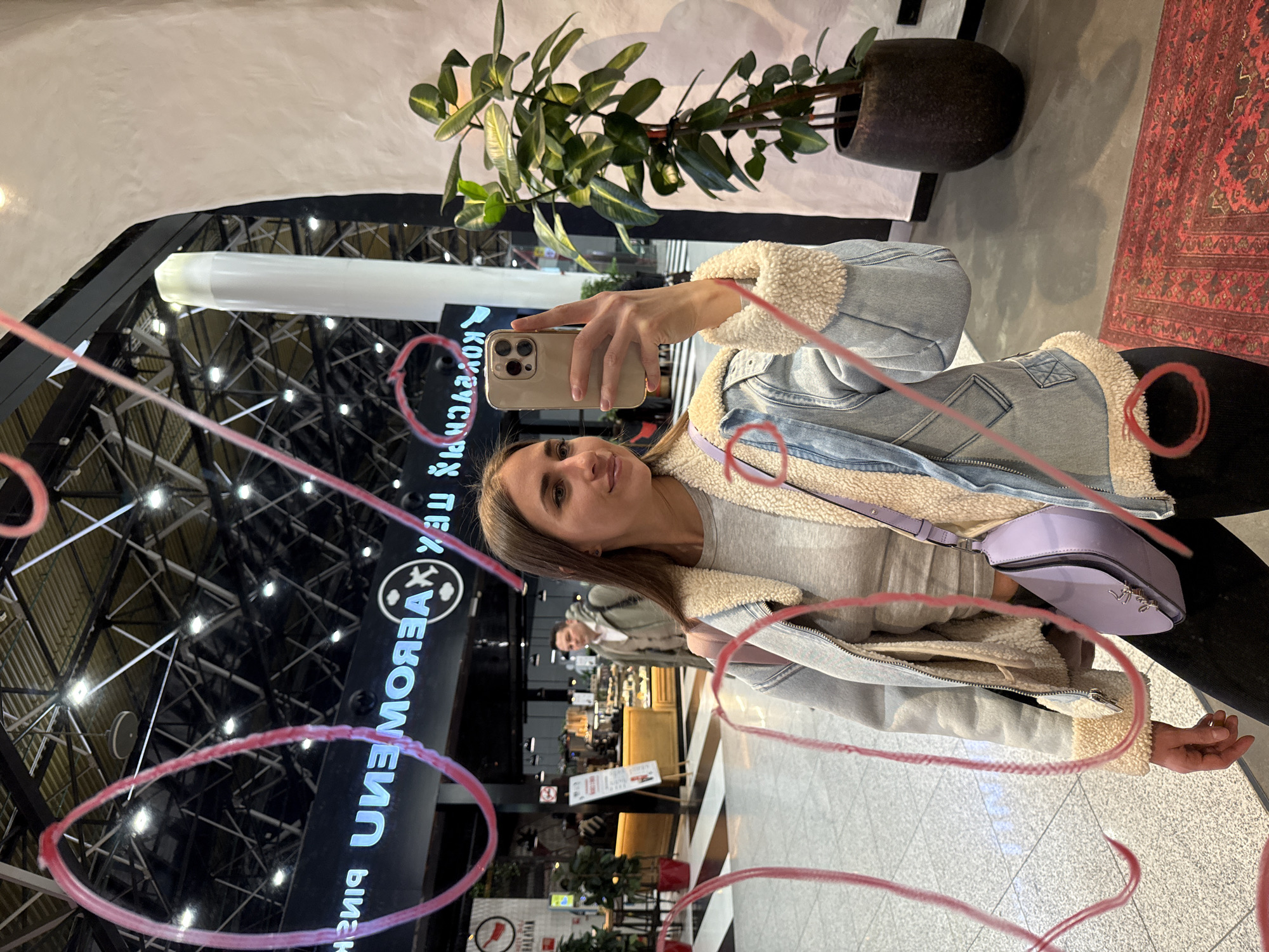A young adult female is depicted taking a selfie in a reflective surface within Sheremetyevo International Airport in Khimki, Russia. The woman, with long brown hair, wears a light-colored long-sleeved top, a light blue denim jacket with cream sherpa lining, black trousers, and a light purple cross-body bag. She holds a light-colored smartphone in her right hand, angled to capture her reflection.
The reflection reveals a large indoor public space consistent with an airport terminal, featuring a high ceiling with exposed dark metal framework and numerous recessed lights. Mirrored white illuminated Russian text "ШЕРЕМЕТЬЕВО" (Sheremetyevo) and an airport symbol (a plane within a circle) are prominently visible on a dark background. Below this, additional mirrored text reads "КОВАСАК ПИХ". Further back in the reflection, a cafe or shop is visible with a wooden counter, display cases containing pastries, and a male individual in a light brown coat walking past. The floor in the reflection is light gray tile with a section of black and white stripes near the cafe.
To the right of the woman, a tall green plant in a dark brown pot stands on a red patterned rug on the floor. A tall white cylindrical pillar is also partially visible on the right. The reflective surface itself is marked with multiple prominent, hand-drawn red circles and squiggles.