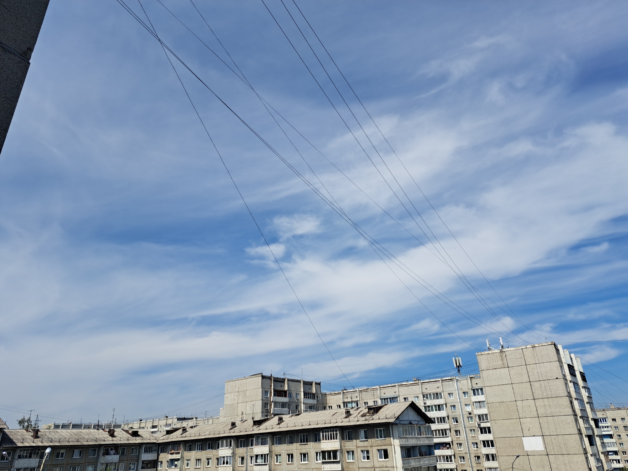 The image shows a view of residential buildings against a cloudy blue sky. There are several power lines running diagonally across the frame. The buildings appear to be apartment blocks, common in many Soviet-era urban areas. The architecture suggests a location in Russia, likely Irkutsk given the provided context. The scene is devoid of people, and no specific actions are depicted. The environment is urban, with the focus on the sky and the residential structures.