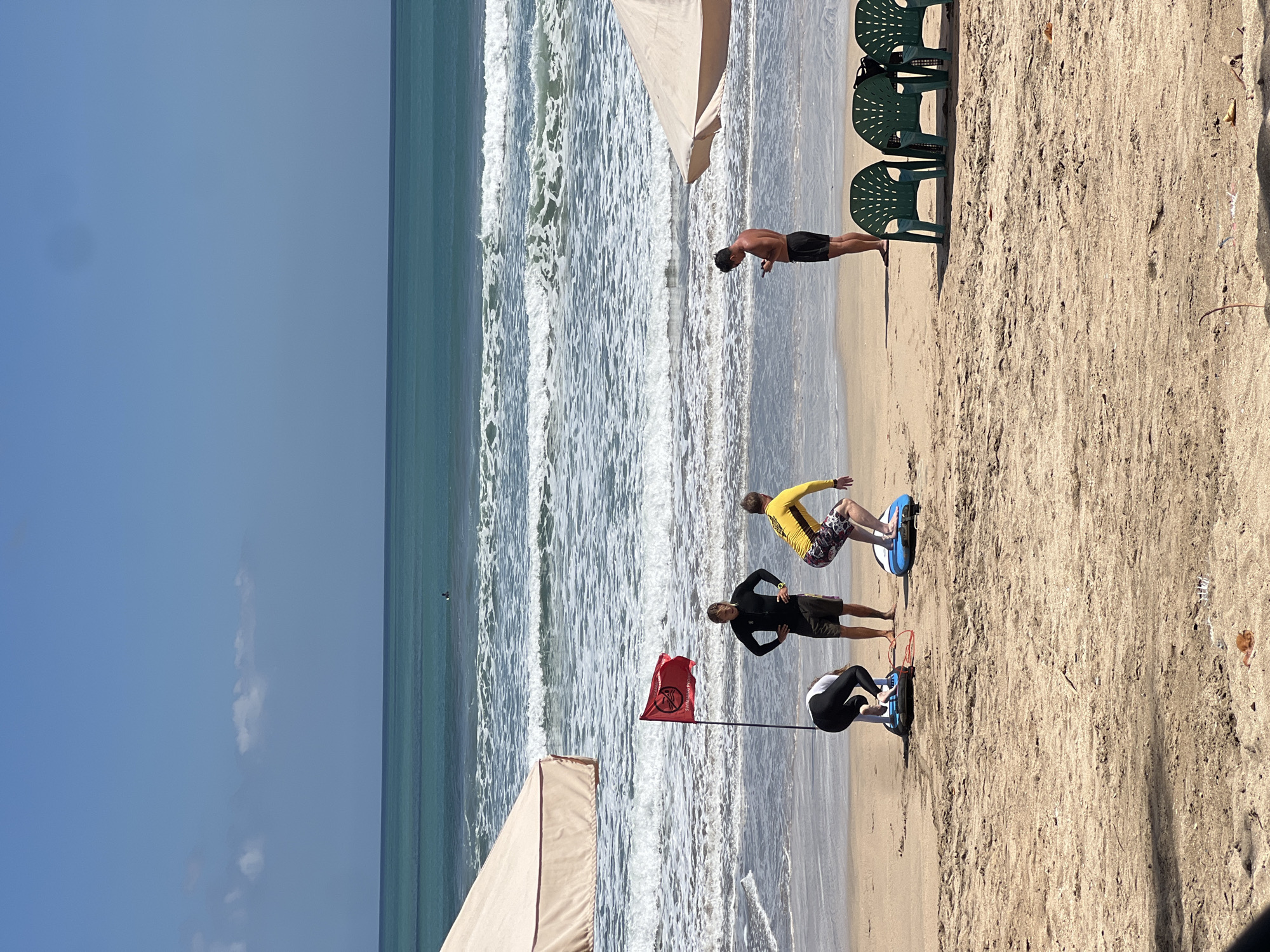 The image depicts a sandy beach with the ocean in the background, likely in Kuta, Indonesia. Several individuals are present on the beach, some engaged in what appears to be surf training. One person in a black wetsuit is standing, observing two others on what look like balance boards. One of these individuals is in a crouched position on a blue board, and the other, wearing a yellow long-sleeved shirt and patterned shorts, is in a surfing stance on a similar board. A red flag with a symbol is planted in the sand near the standing individual. In the distance, a lone surfer can be seen in the ocean, and further out, the turquoise waves crash on the shore under a clear blue sky. To the right, a person in black shorts is looking at their phone, and behind them are several green plastic beach chairs. Two large, light-colored beach umbrellas are partially visible on the left and right sides of the image, suggesting a developed tourist area. The sand is textured with debris, including dried leaves and small twigs.