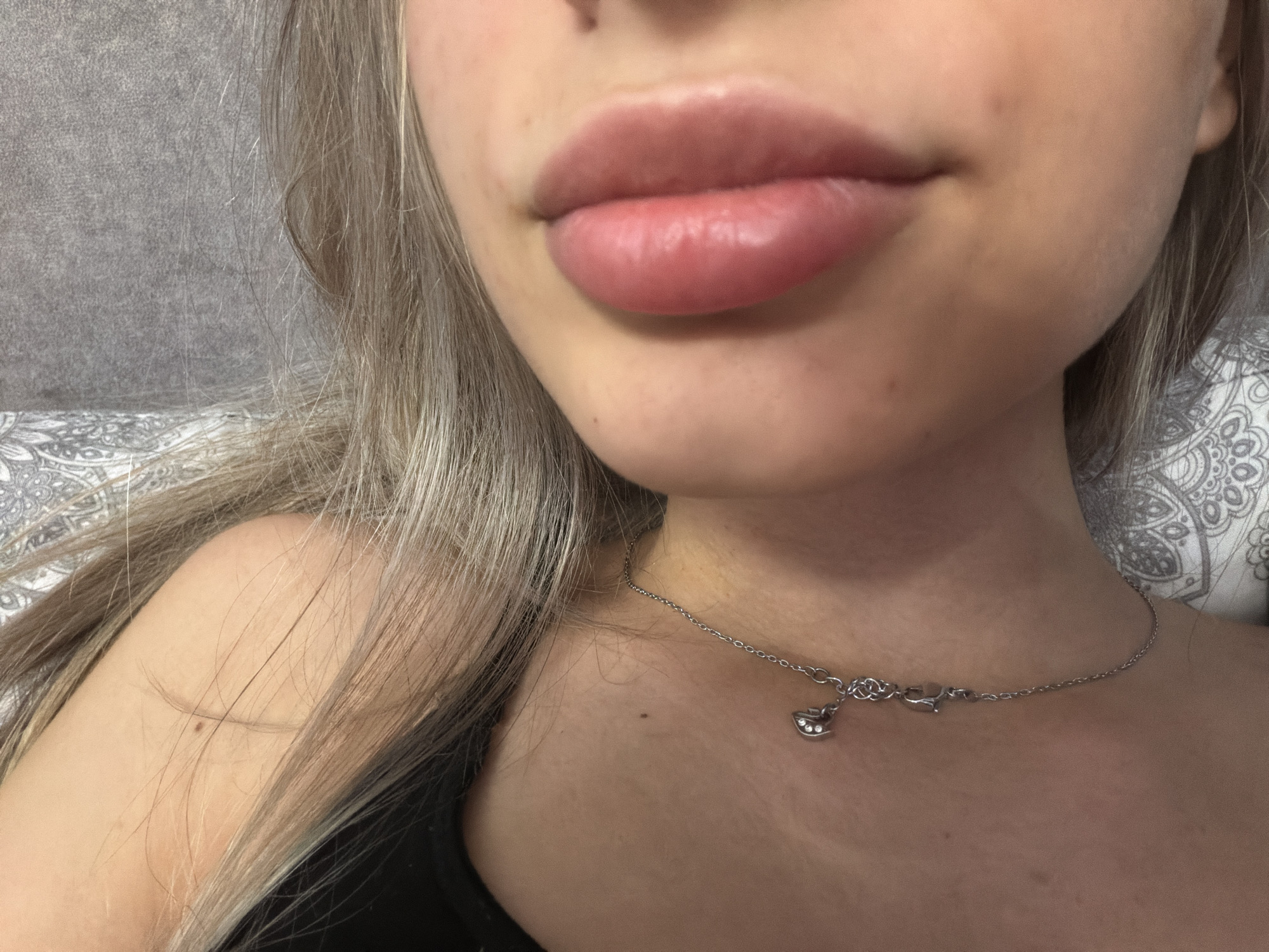 The image is a close-up, head-and-shoulders shot of a young woman. She has fair skin, blonde hair, and prominent, full lips. She is wearing a dark-colored top and a delicate silver necklace with a small charm. Her hair is partially covering her face and shoulders. The background is out of focus and appears to be a textured surface, possibly a wall or fabric, with a hint of a patterned textile on the right. The lighting suggests an indoor setting, possibly natural light from a window or soft artificial light. The image quality is good, with a shallow depth of field that blurs the background and draws attention to the subject's face and neck. There is no indication of the location being in the city of Ruse, Bulgaria.