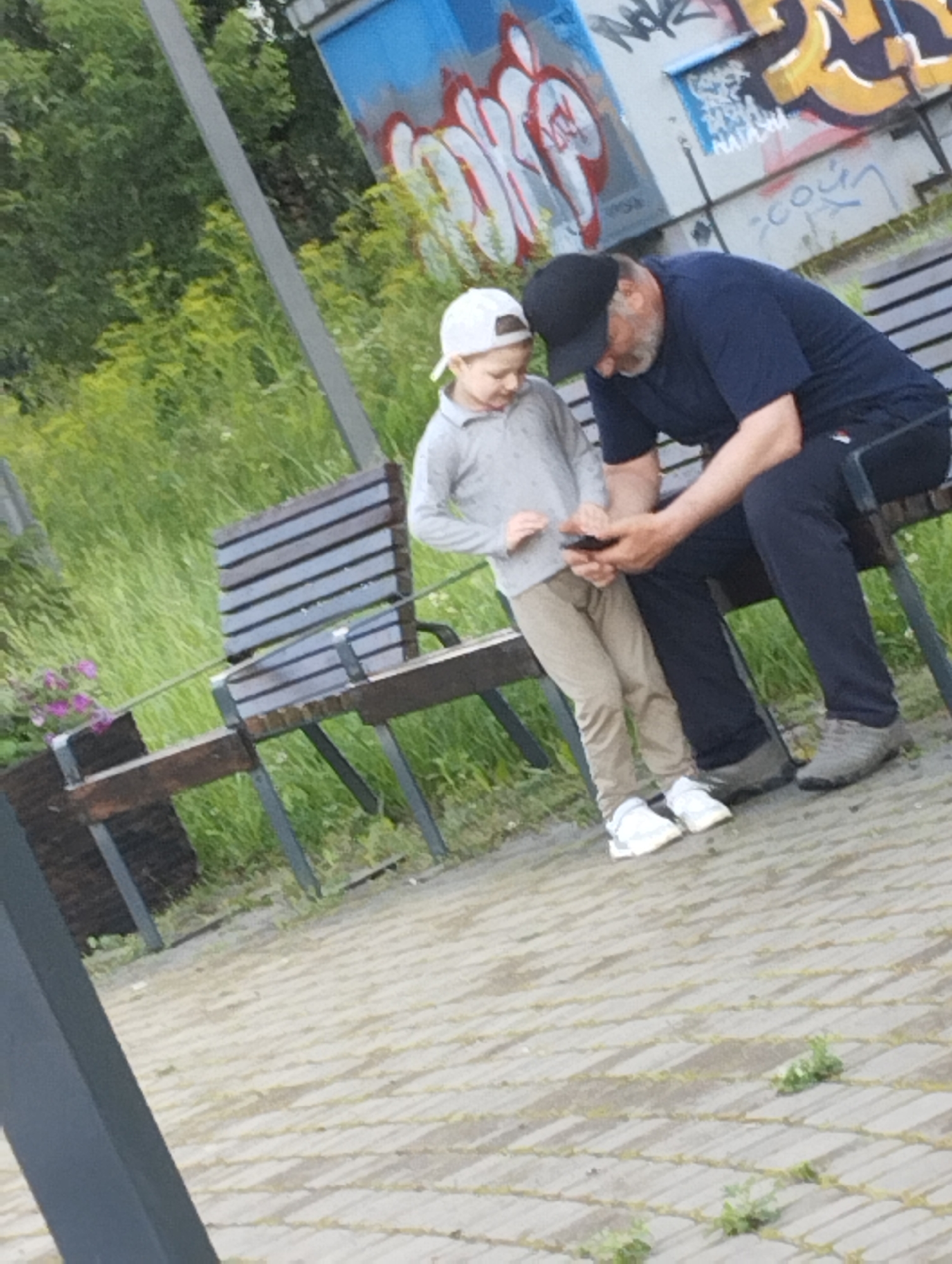Two individuals, a man and a child, are seated on a wooden bench. The man is wearing a dark blue t-shirt, dark trousers, and a black baseball cap. The child is wearing a light grey long-sleeved shirt, light brown trousers, and a white baseball cap. The man is holding a smartphone in his right hand, and the child is looking at the phone. The bench is situated outdoors, with a paved ground and lush green grass and trees in the background. A graffiti-covered structure is visible in the background. The scene is likely located in Kingisepp, Russia, as indicated by the prompt, potentially in a park or public area.