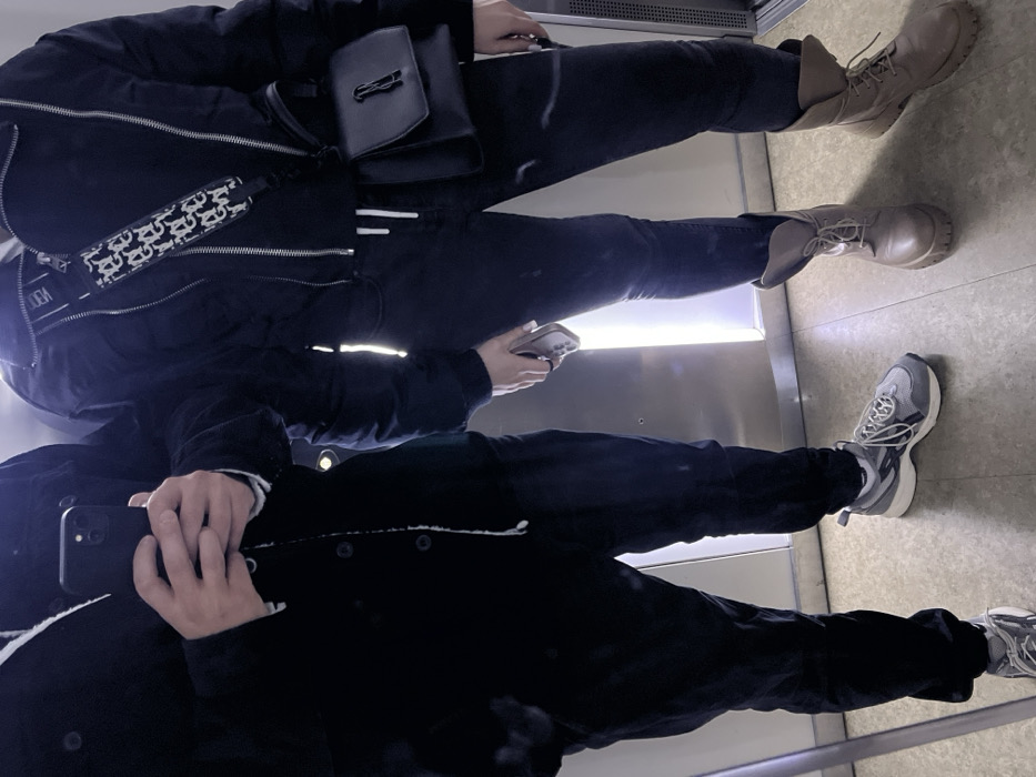 Two people stand in an elevator. The person at the top wears a black jacket with a Dior crossbody bag, dark jeans, and tan combat boots. They hold a phone. Reflected below, the second person wears a black jacket with a white lining, black pants, and gray sneakers. They are holding a phone. The elevator walls are metal and contain a light.