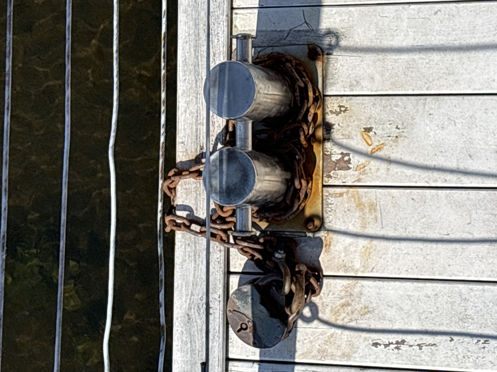 The image shows a close-up, overhead view of a wooden dock on the Moskva River in Zvenigorod, Russia. The dock is constructed from weathered, white-painted wooden planks. On the dock, there are two shiny, cylindrical bollards, likely made of metal, positioned side-by-side. A thick, rusty chain is wrapped around the base of the bollards, connecting them and also extending to a large, circular, rusty lock on the left side of the image. The lock is attached to another chain loop. The water of the river is visible in the background, appearing dark and reflecting thin, horizontal lines of light, possibly from overhead structures or the sun. The overall scene depicts elements of a functional marine environment, with the dock, bollards, and chains suggesting a place where boats are secured.