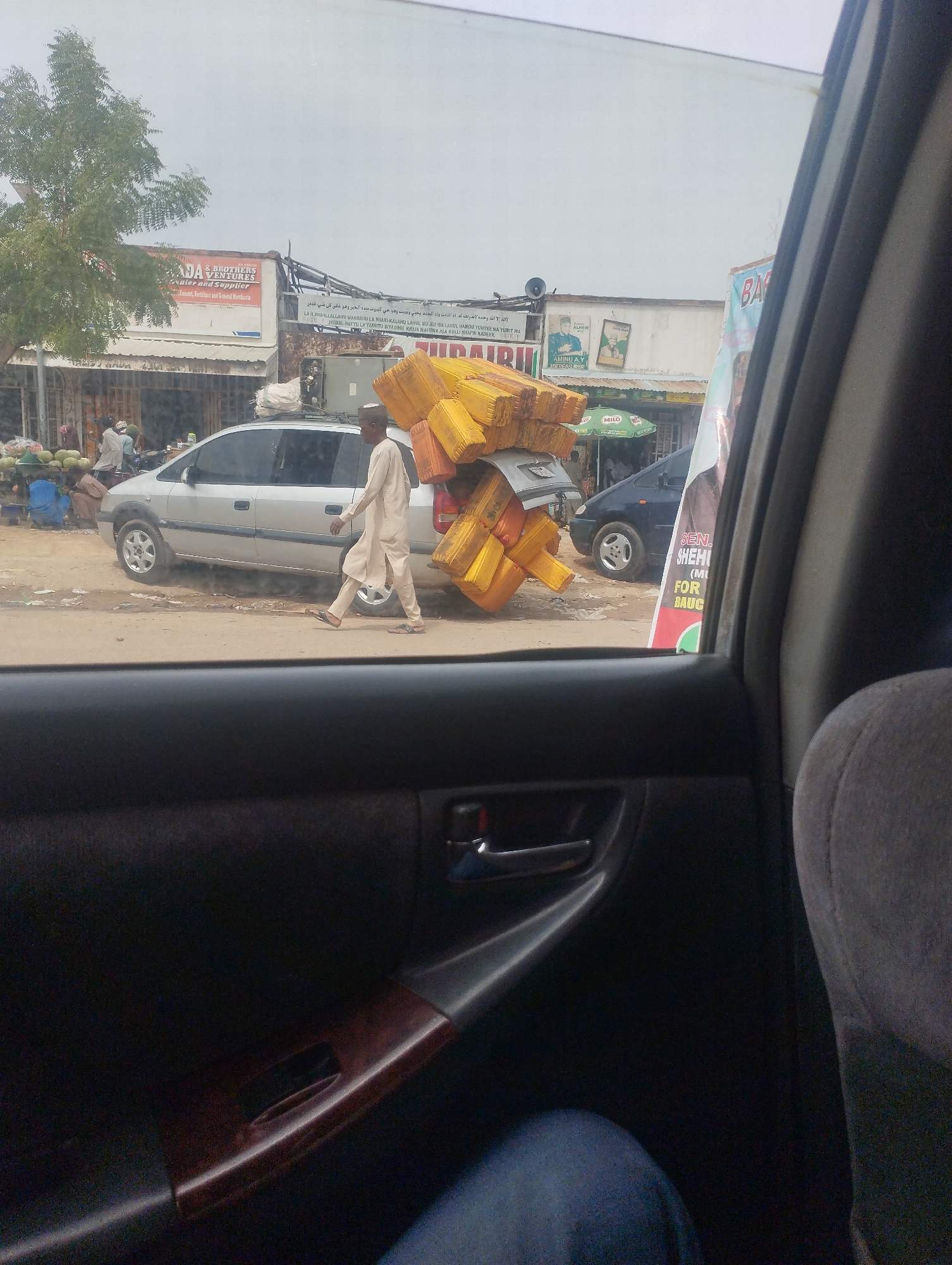 The image shows a scene in what appears to be Bauchi, Nigeria, taken from inside a vehicle, focusing on the street activity visible through the car window. A man dressed in a light-colored robe and a small cap is carrying a large load of yellow jerrycans on his back as he walks past a silver minivan. To the left, there are vendors selling watermelons. Buildings line the street, with signs displaying business names like "ADA & BROTHERS VENTURES" and political posters with the name "SEN. SHEHI (MI FOR BAUC)." A Milo umbrella is visible in front of one of the shops. The scene depicts a typical urban street with commercial activity, people engaged in trade, and the everyday transport of goods.
