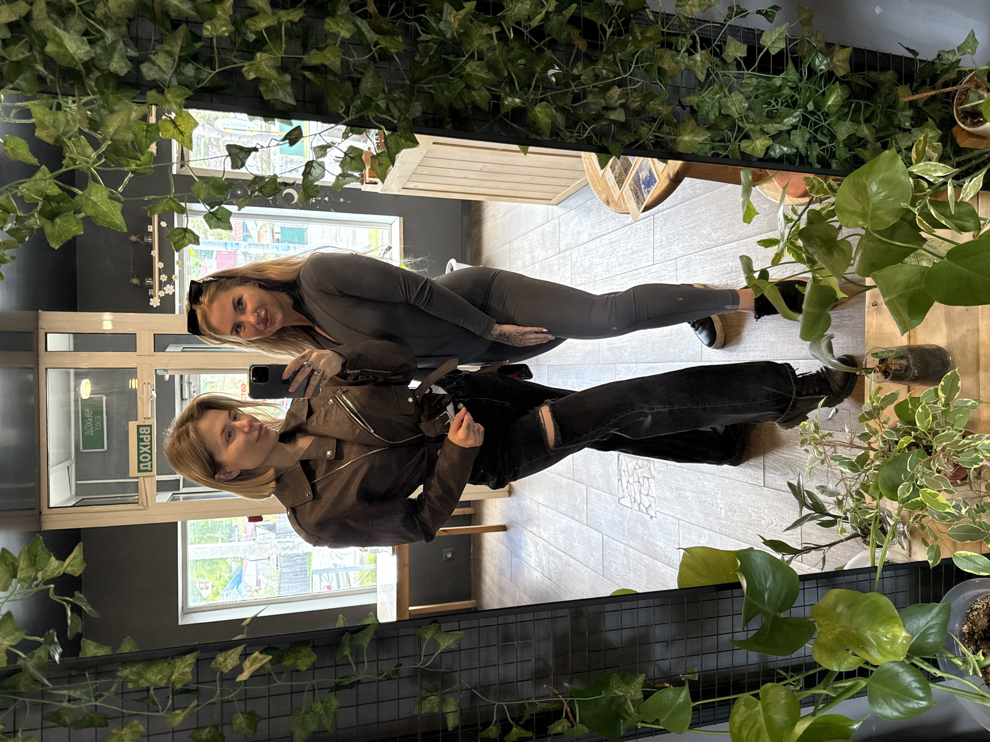 Two women are taking a mirror selfie indoors, likely in a commercial establishment, with the reflection showing elements of the location and the women themselves. One woman, visible in the lower half of the image, is wearing a brown jacket and black jeans with a rip in the knee. She is holding a mobile phone, presumably to take the photo. The other woman, positioned in the upper half of the image, is wearing a grey bodysuit and is smiling at the camera. The location appears to be in Mayma, Russia, as evidenced by the reflected signs with Cyrillic text indicating "EXIT." The interior features neutral grey walls, light-colored flooring, and windows that offer glimpses of the outside. A plant is placed in front of the mirror on the bottom and top, creating a frame around the reflection.