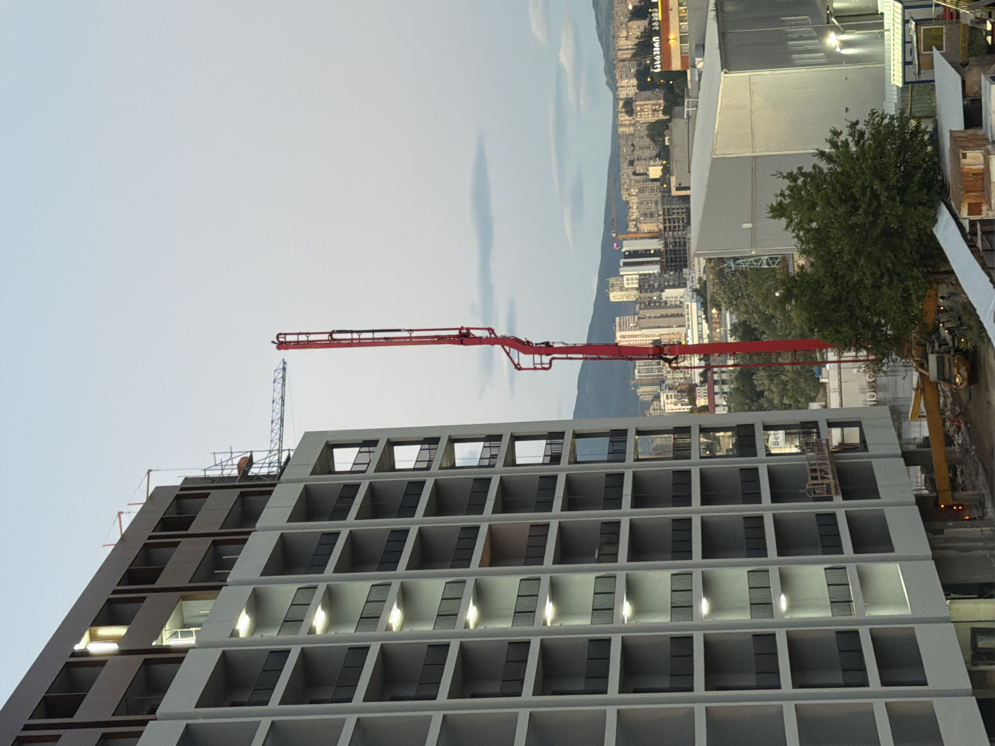 An outdoor, medium shot captures a modern, unfinished apartment building under construction in Tbilisi, Georgia, against a pale blue, cloudy sky. The building, predominantly gray with dark brown accents, features multiple floors with visible balconies or open-air spaces. Individual balconies are equipped with dark railing. Lights are visible in some of the interior spaces on the lower floors. To the right of the apartment building, a tall, red concrete pump boom is prominently positioned. In the background, the cityscape of Tbilisi unfolds, showcasing a dense collection of apartment buildings and other structures stretching towards distant, hazy mountains. A construction crane is also visible in the mid-ground. The lower portion of the image reveals construction materials and vehicles, including a yellow crane arm. The overall scene depicts an active construction site in an urban environment.