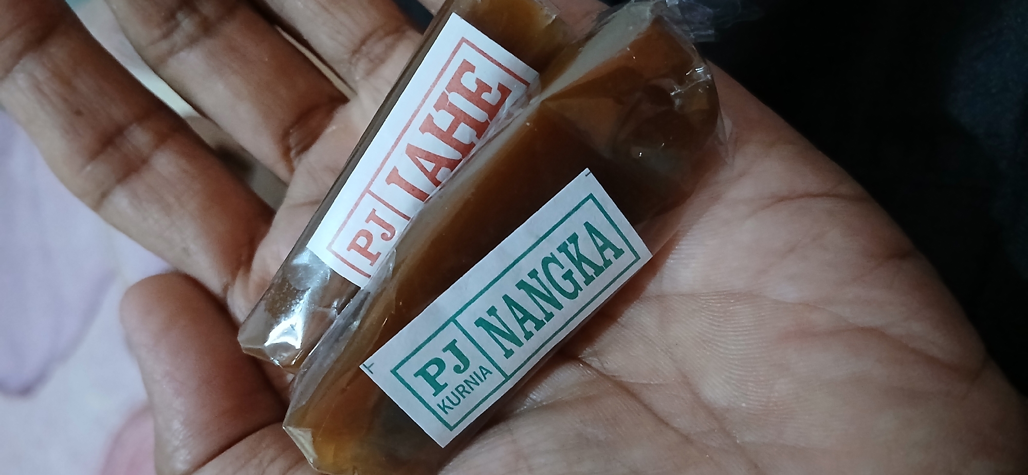 Two individually wrapped candies are held in a person's hand. The candies are rectangular and appear to be a golden-brown color. Each candy is wrapped in clear plastic and has a white label with text on it. The label on the candy in the foreground is green with the text "PJ NANGKA" and "KURNIA" in a smaller font. The label on the candy in the background is red with the text "PJ JAHE". The background of the image is out of focus and shows a light-colored fabric, possibly a tablecloth or clothing. The image was taken at an unspecified location in Samarinda, Indonesia.
