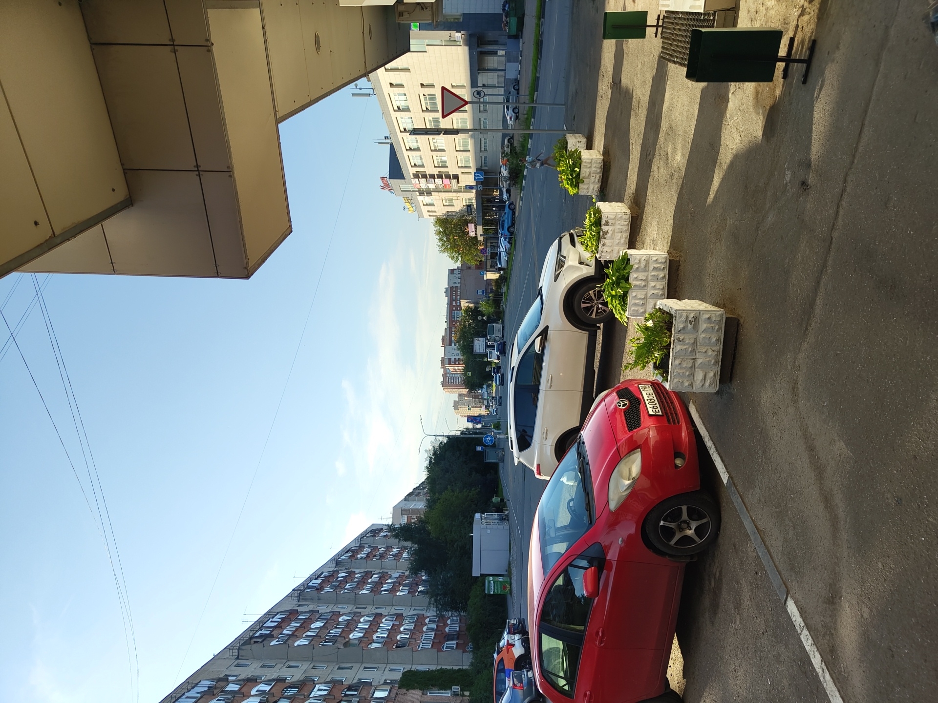 The image depicts a street scene in Zhukovsky, Russia, with a view of a long, multi-story apartment building on the left and a modern building on the right. In the foreground, a red hatchback is parked, and behind it, a white SUV is also parked. Several rectangular planters with greenery are placed along the sidewalk. The street in the background shows traffic with cars and a bus. The sky is clear blue with some thin power lines visible. The architecture of the buildings suggests a post-Soviet urban environment. There are no prominent actions or interactions visible, and the scene appears to be a typical daytime street view.