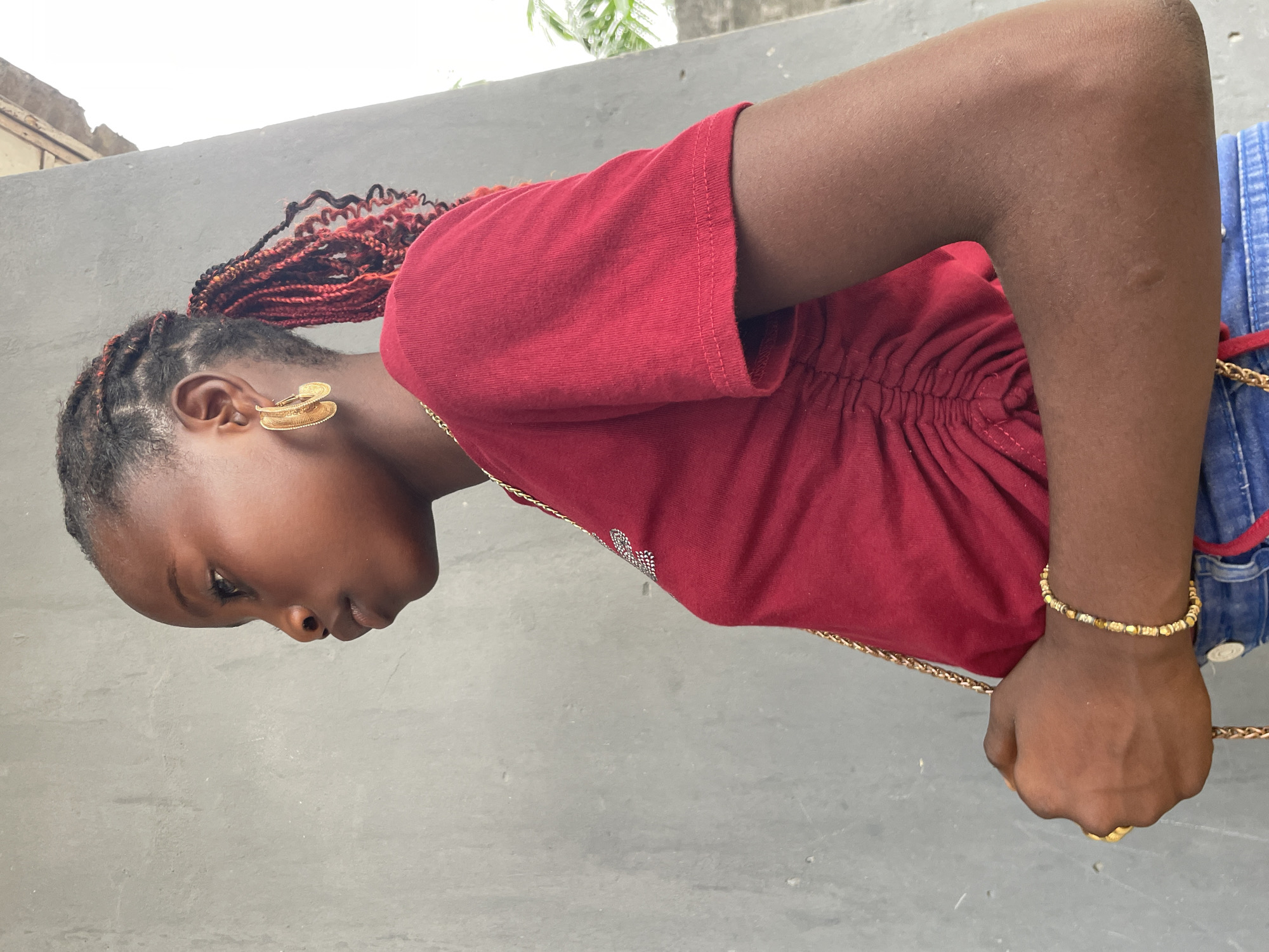 An outdoor, profile shot depicts a young dark-skinned individual, likely female, from the chest up. She is presented with her head turned to the left and tilted downwards, showcasing a neutral expression. Her hair is styled in intricate reddish-brown braids, swept upwards.
She wears a short-sleeved top in a rich red or maroon color, featuring ruched detailing along the side and subtle silver-colored embellishments on the front. Her attire includes blue denim bottoms, partially visible. Prominent gold-colored accessories adorn her: large, intricately designed hoop earrings, a thin chain necklace, a beaded bracelet on her left wrist, and a ring on one finger of her left hand. This hand is shown holding a gold-colored chain, possibly a strap from a bag. A small nose ring is also visible.
The background consists of a plain, light grey concrete wall. Above the wall, glimpses of green foliage and a bright sky indicate an outdoor setting during daytime, with clear, even lighting. The overall scene suggests an urban or semi-urban environment, consistent with locations in Badagry, Nigeria. There is no visible text in the image.
