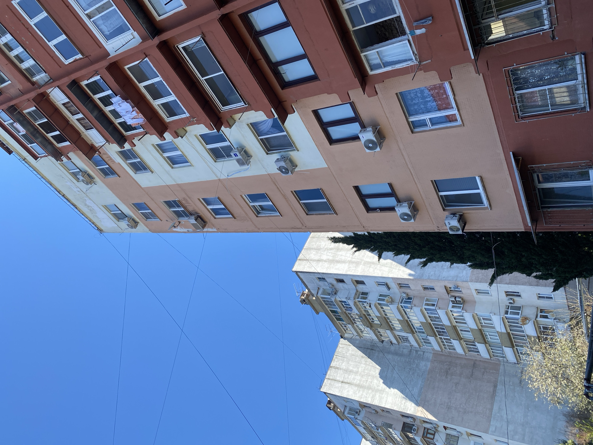 The image is a low-angle shot of two buildings against a clear blue sky. The top building is a multi-story structure with a color scheme of brown, beige, and white. Numerous windows are visible, some with air conditioning units mounted below. Clothes are hanging out of a window. The bottom building is a gray and white multi-story structure, with a similar arrangement of windows and air conditioning units. Trees are visible between the buildings. Thin wires are strung across the sky between the buildings. The image appears to be taken in Sochi, Russia, based on the architectural style.