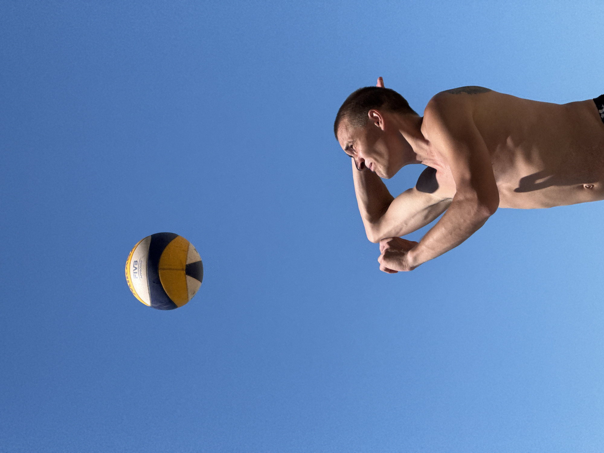 In the city of Nebug, Russia, under a clear blue sky, a shirtless man with short brown hair is looking up at a volleyball. He is in the process of playing volleyball, with his arms raised and hands positioned as if to hit the ball. The volleyball is in the air above him. The man is wearing black swim trunks.