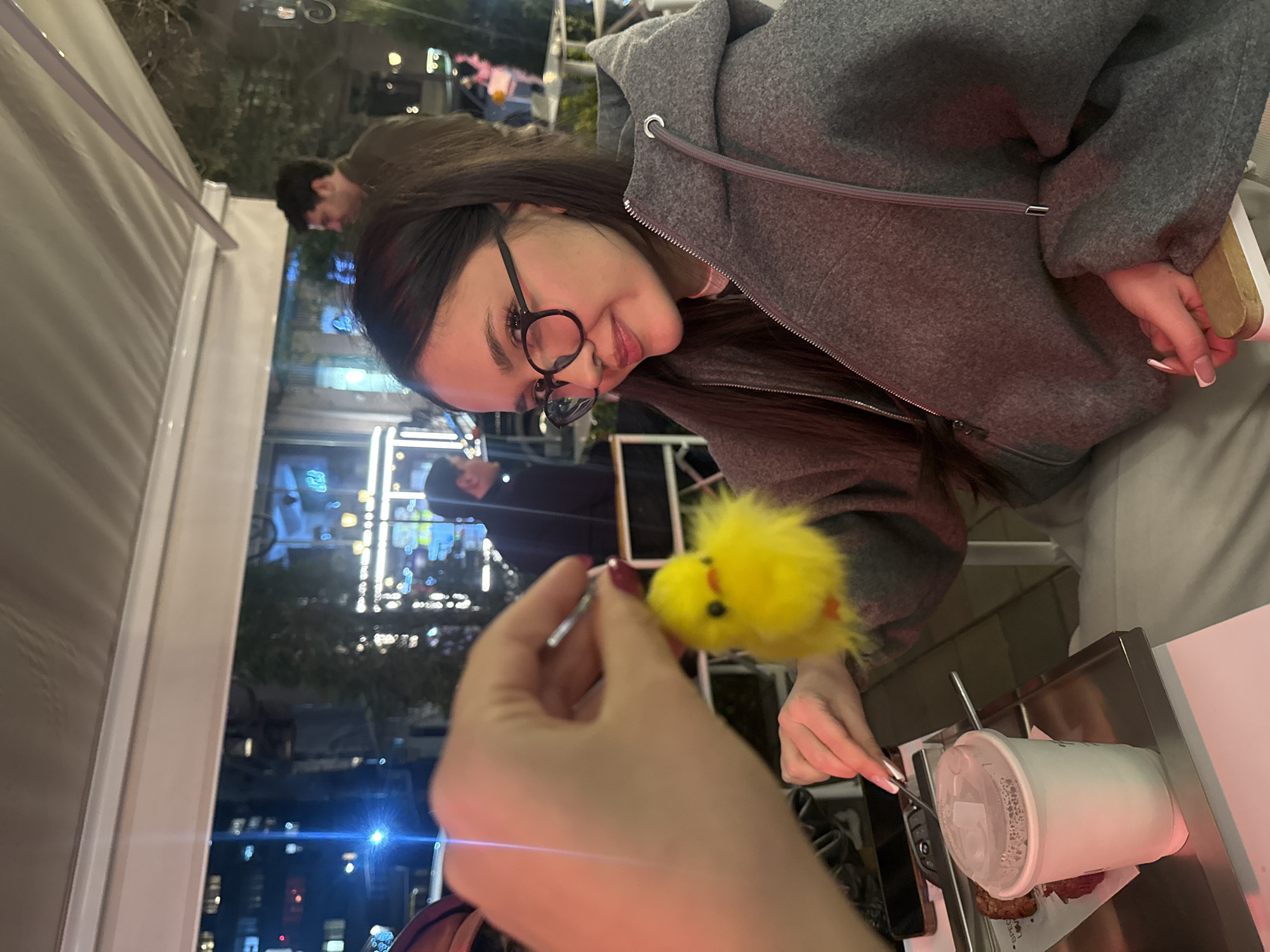 The image shows a woman wearing a gray hoodie and glasses, seated at an outdoor table. Another person is holding a small, yellow, fuzzy chick toy on a stick. The woman is smiling and looking at the toy. On the table, there is a metal tray with a white cup, a utensil, and what appears to be food. The background shows a nighttime urban environment with buildings and streetlights, which is identified as Yerevan, Armenia, from the bright neon signs. Other people are visible in the background, indicating a public space.