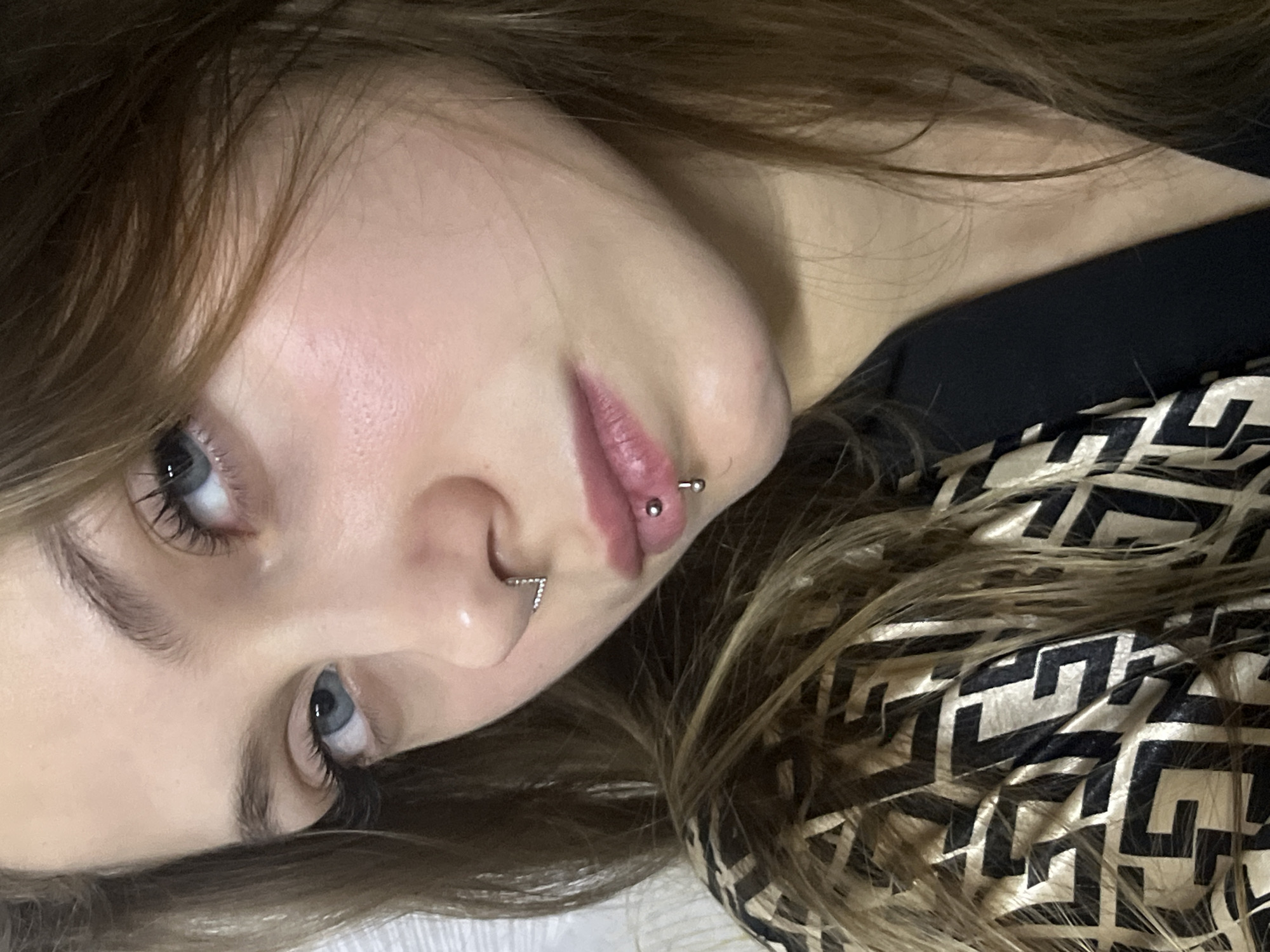 A close-up, angled shot focuses on a woman's face. Her skin appears smooth, and her light-colored eyes are visible. She has piercings in her nose and lip. Her brown hair is styled loosely around her face. A patterned fabric, possibly clothing or a blanket, is partially visible in the lower right corner of the frame. There is no indication of the location of Фоки, Russia.