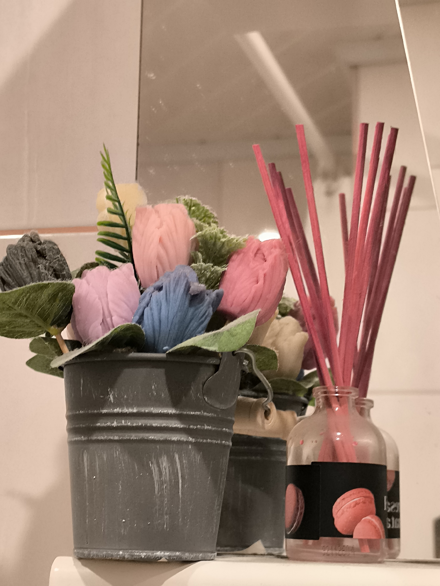 A close-up shot captures a decorative arrangement on a shelf. In the foreground, a grey, distressed metal bucket holds an assortment of artificial flowers in pastel shades of pink, blue, and yellow, accented with faux greenery. To the right, a glass diffuser bottle with pink reeds sits next to a blurred reflection of the flowers. A black label on the diffuser features images of macarons. The background is a reflective surface, likely a mirror, showing a light-colored wall and a hint of a doorway or corner. No people are present, and no identifiable location in Klaipėda, Lithuania is depicted.