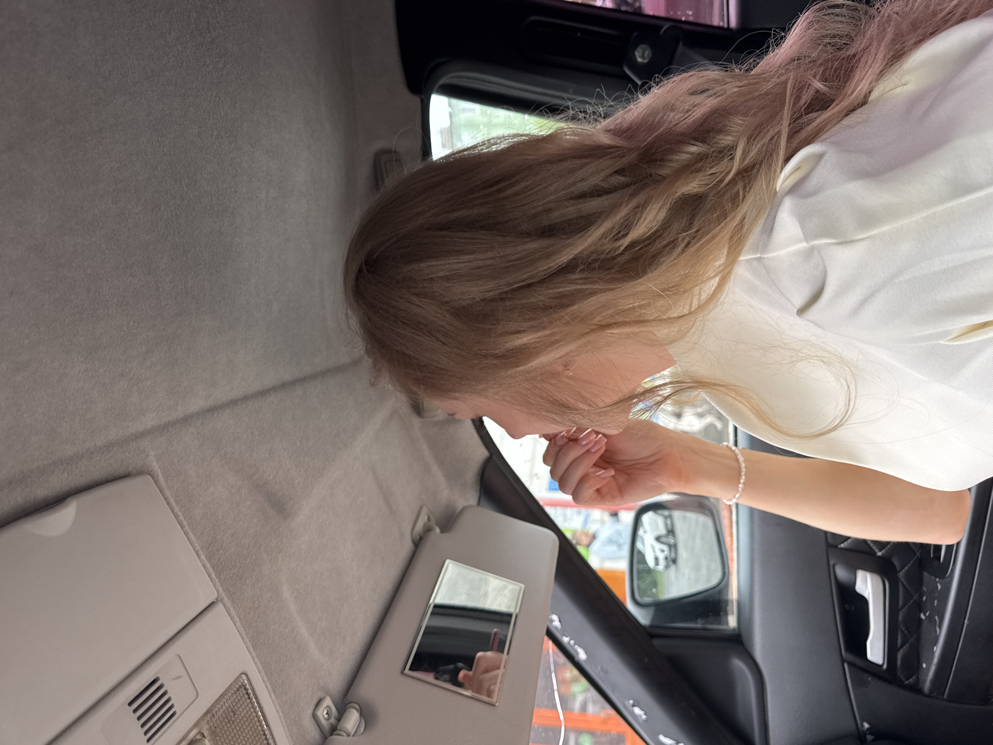 In a vehicle, a young woman with blonde, wavy hair is seen from the side, looking into a visor mirror and applying makeup. She is wearing a white top and a pearl bracelet on her left wrist. The car's interior is visible, including the ceiling, sun visor with a mirror, side door panel, and a side mirror reflecting a white car and a person in the background. The scene is consistent with a street or parking area in Perm, Russia, possibly near a car wash or commercial establishment.