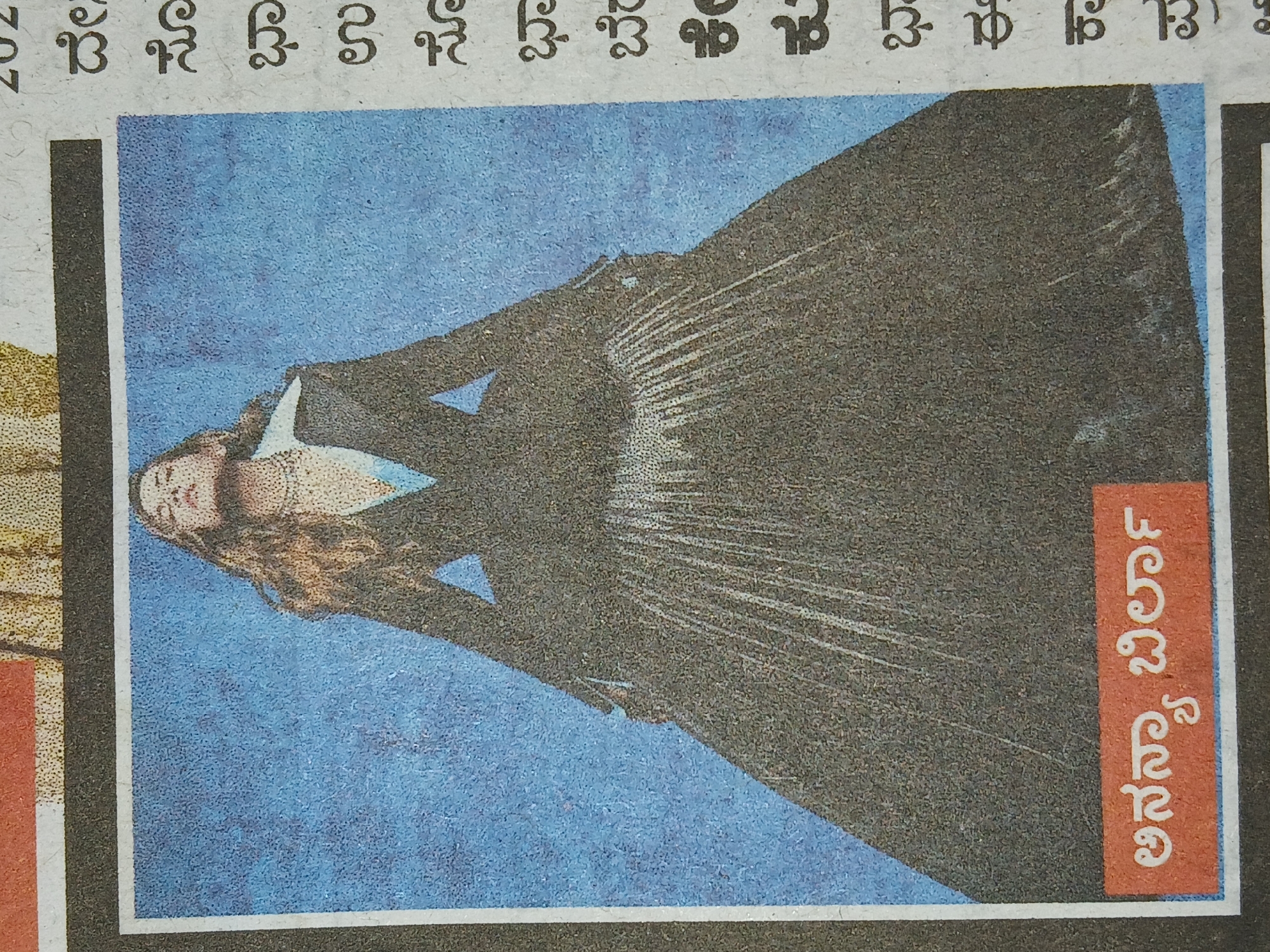 A photograph of a newspaper clipping features a full-length image of a light-skinned woman with long, wavy brown hair, wearing a black outfit that appears to be a dress or a combination of top and skirt. She is leaning back at an angle, with her face and upper body visible, set against a solid blue background. The newspaper clipping has a black border with a thin white line inside. To the right of the image is text in Kannada script on a red rectangular background. The photograph is taken in a setting where the newspaper print is slightly visible in the background, suggesting the location is Bengaluru, India. The image appears to be a promotional or advertisement piece.