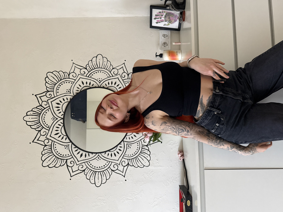 An indoor scene in Rostov-on-Don, Russia, depicts a female subject with long, bright red hair, standing relaxed with eyes closed and a slight smile, head tilted back and to the left. She wears a black ribbed crop top, dark high-waisted jeans, a silver necklace with a cross pendant, a silver bracelet on her left wrist, and a septum nose piercing. Her arms are extensively tattooed with intricate designs, as is her lower abdomen below the crop top. She is positioned in front of a light-colored, textured wall adorned with a prominent circular mirror surrounded by a black mandala-style decal. Beneath the mirror, a white dresser or chest of drawers holds various objects: a dark-colored box with a red lining, a pink scrunchie, a single pink flower (possibly a tulip), a white pump bottle with an orange label, a small pink cylindrical object, a white double electrical outlet, a framed picture of a colorful handprint labeled "FAMILY," and a black object with metallic accents.