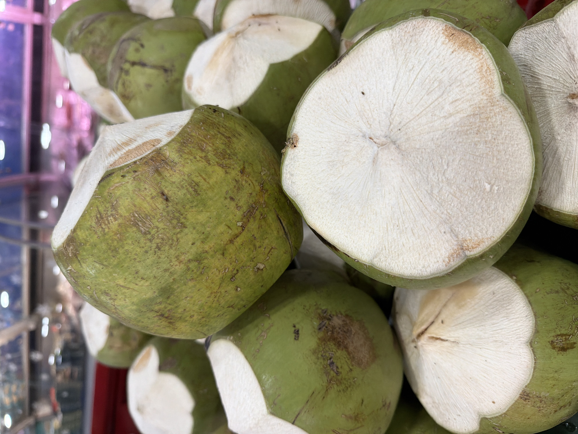 The image shows a pile of freshly prepared coconuts for sale in a market in Nha Trang Ward, Vietnam. The coconuts are green and have been partially peeled, exposing the white flesh inside. Several coconuts are stacked on top of each other, with some cut in half to reveal the interior. The background is blurred but appears to be a marketplace with shelves and lighting. There are no people or animals visible in the image.