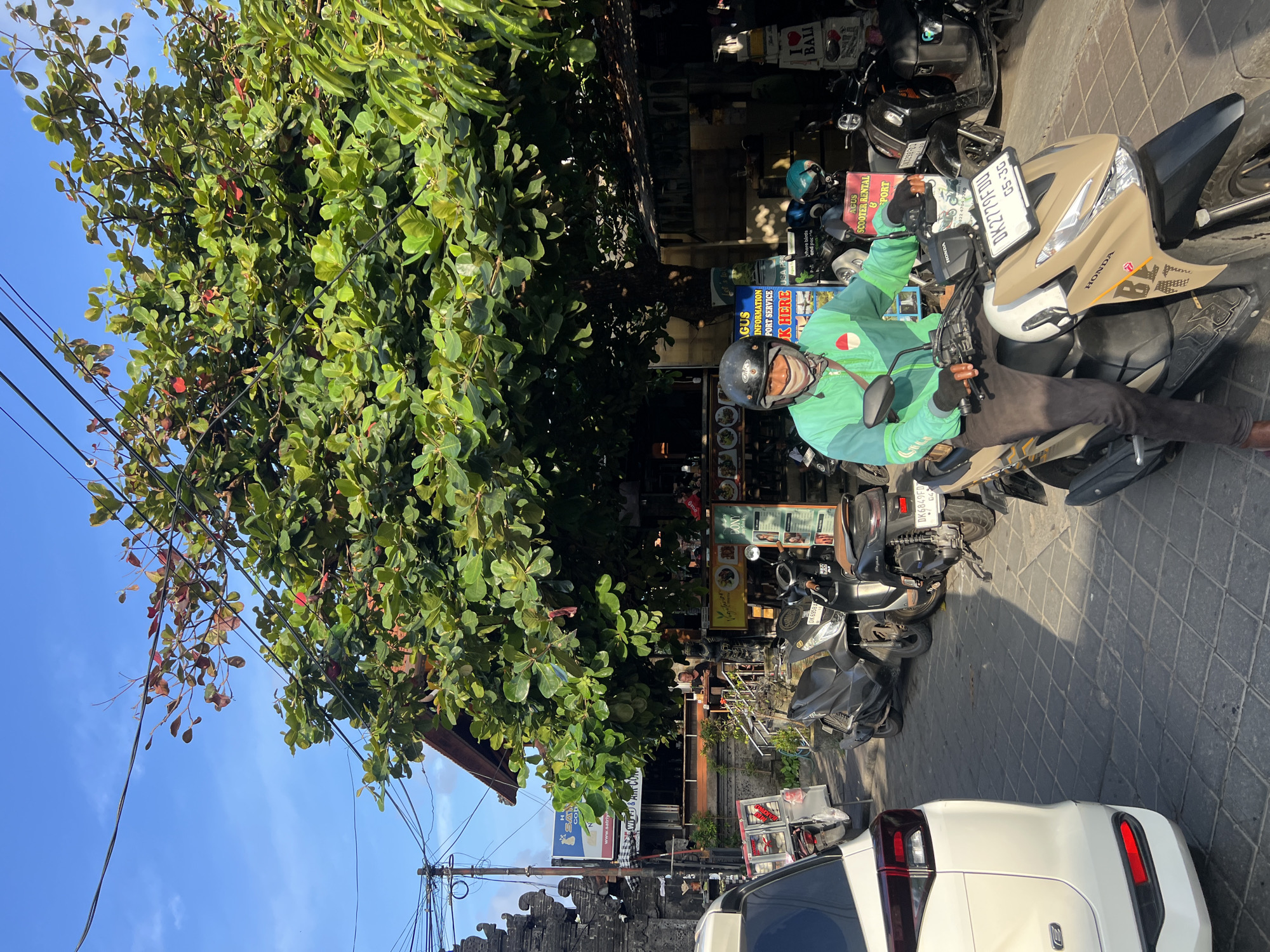 In Kuta, Indonesia, under a tree with large green leaves and some red accents, a person wearing a light green jacket, black helmet, and protective mask rides a beige and black scooter. The scooter has a license plate that reads "DK 2779 FDU". The individual is wearing dark pants and black gloves, and their right arm is resting on the handlebars. The scooter is parked on a stone-paved street, with other scooters visible in the background. To the left of the scooter, a white SUV is partially visible. Buildings with traditional Balinese architecture and shops line the street, with signage indicating services like "AGUS SCOOTER RENTAL" and "I LOVE BALI". Power lines are visible above the scene. The sky is clear and blue, suggesting a sunny day.