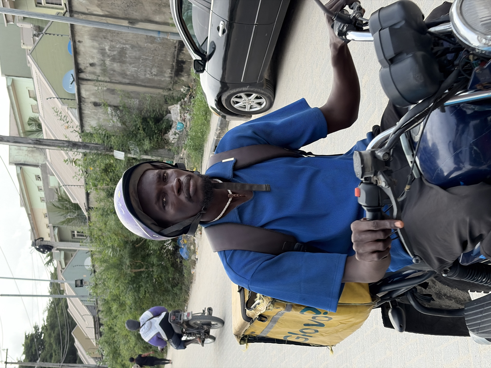 A dark-skinned man wearing a purple and white helmet, a blue collared shirt, and dark pants is riding a motorcycle. He has a brown backpack on and a yellow delivery bag with "Glovo" written on it is attached to the back of the motorcycle. The motorcycle is dark blue. In the background, there is a black car, a concrete wall with graffiti, lush green bushes, and several houses with light-colored roofs. A person in a purple and white jacket is riding another motorcycle in the distance. The scene is located on a paved road in Igboefon, Nigeria.