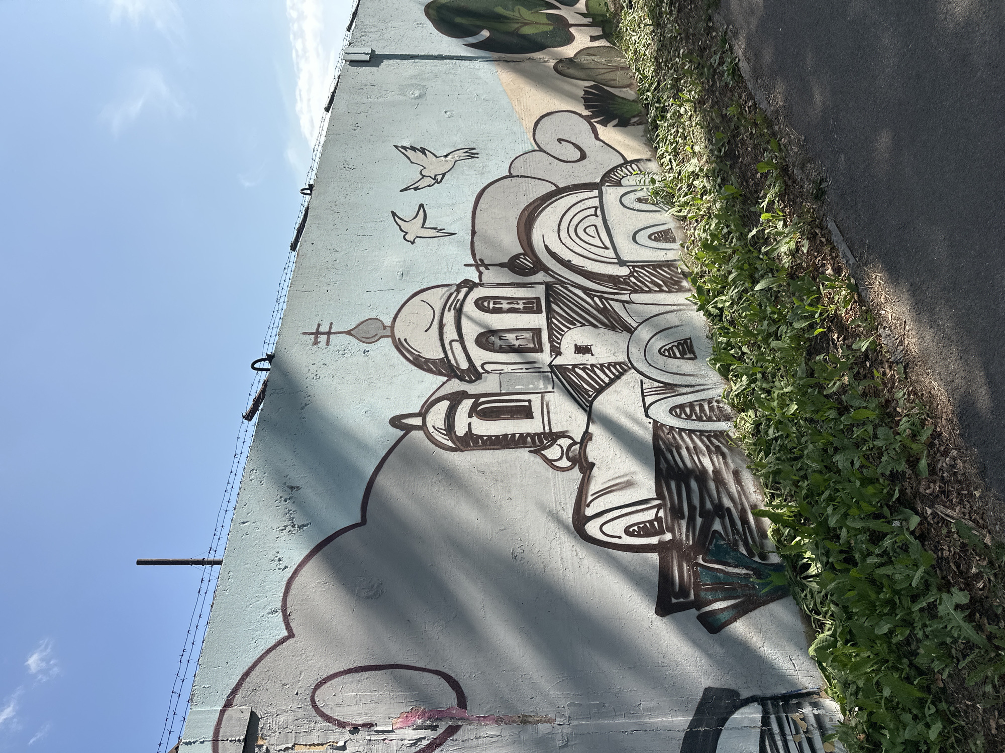 A sunlit, outdoor scene captures a light blue concrete wall adorned with graffiti art, located in Biysk, Russia. The graffiti depicts a stylized, outlined church with domes and crosses, set against a backdrop of outlined clouds. Two birds are painted in flight above the church. To the right, there are outlines of trees. Barbed wire runs along the top edge of the wall, and a slender, dark pipe extends upwards from behind it. Shadows from an unseen object cast diagonal lines across the wall. The base of the wall is bordered by a strip of overgrown green vegetation and a paved pathway. The sky is clear and blue with a few wisps of clouds.