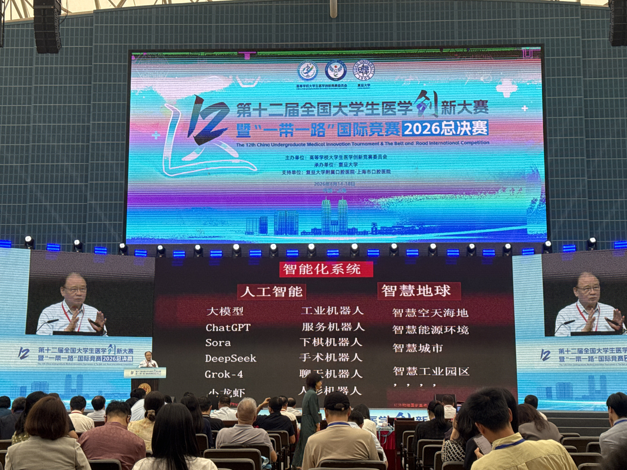 The image shows a wide screen displaying an event titled "The 12th China Undergraduate Medical Innovation Tournament & The Belt and Road International Competition 2026 Finals". The screen is divided into sections with text and graphics. On the left side of the screen, there's an image of an older Asian man speaking at a podium, with a microphone in front of him. The main screen displays various categories related to intelligent systems, including "Artificial Intelligence" and "Smart Earth". Under Artificial Intelligence, there are subcategories like "Large Model", "ChatGPT", "Sora", "DeepSeek", and "Grok-4". Under Smart Earth, there are categories such as "Smart Sky and Sea", "Smart Energy and Environment", "Smart City", and "Smart Industrial Parks". The background of the screen features a colorful, abstract design with a cityscape graphic at the bottom. In the foreground, a crowd of people are seated, facing the screen, suggesting they are attendees at an event or presentation. The overall environment appears to be an auditorium or conference hall. The text on the screen indicates the event is in Yangpu District, China, likely at a university or convention center, given the academic and competitive nature of the event.