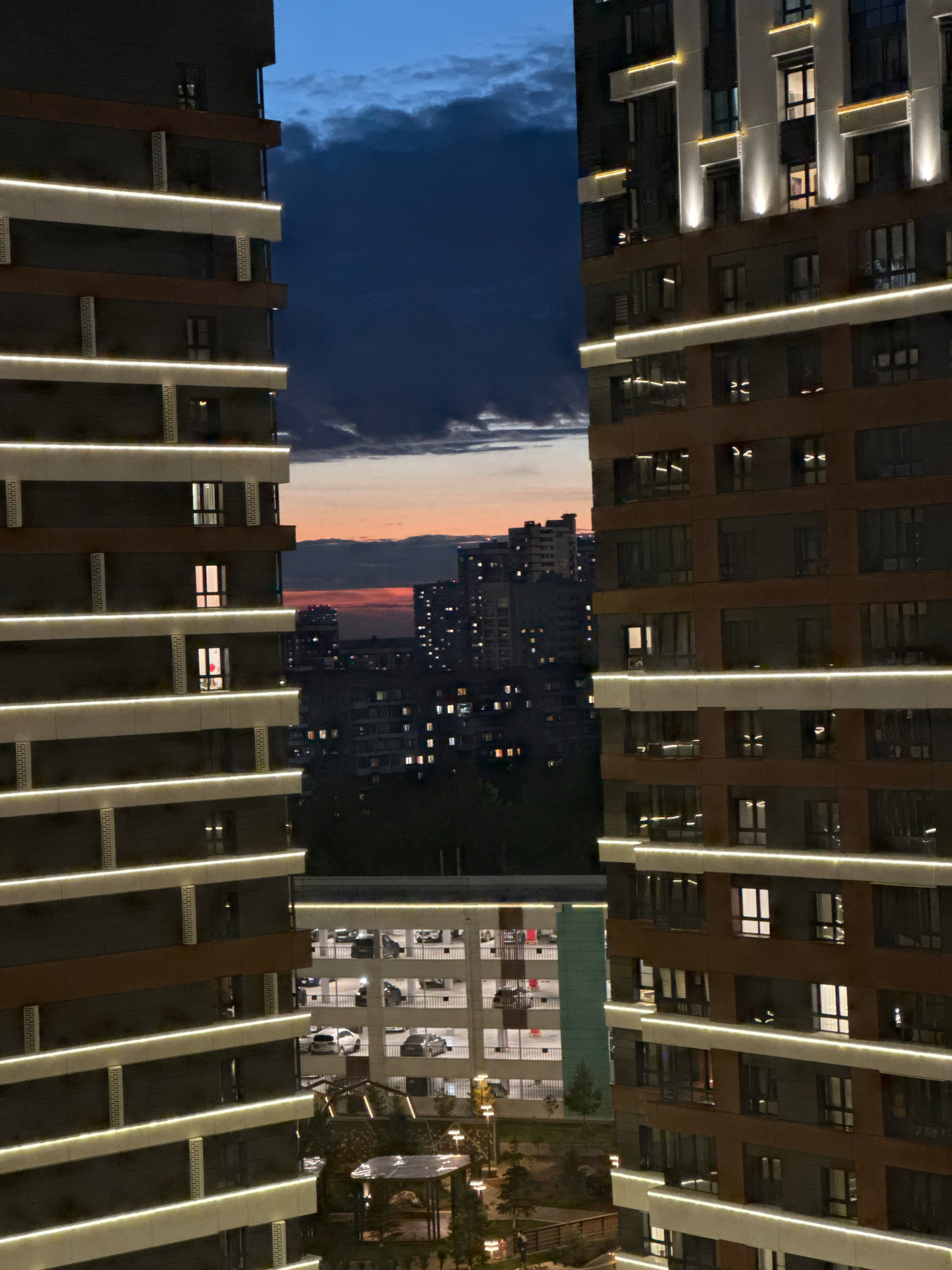 The image depicts a view of a modern residential complex at dusk or twilight, likely in the city of Novosibirsk, Russia. Two tall, multi-story buildings with dark facades and illuminated horizontal bands of light are prominently featured on either side, framing a view of the cityscape beyond. The sky displays a gradient of deep blue at the top, transitioning to orange and pink hues near the horizon, indicating the setting sun.
In the middle ground, several other apartment buildings are visible, with many windows illuminated, suggesting occupancy. A multi-level parking garage with cars parked on its open-air floors is situated between the foreground buildings. The lower portions of the image reveal some landscaping, including trees and what appears to be a small gazebo or sheltered seating area. The overall scene conveys a sense of urban living during the evening hours. No people are clearly discernible.