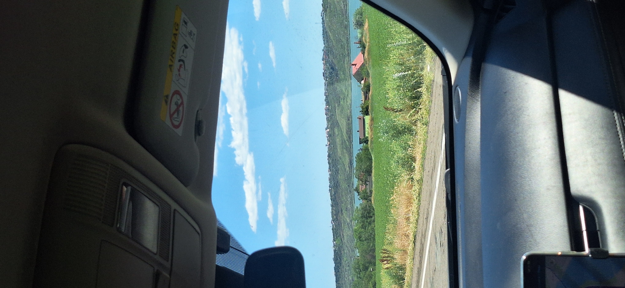 A view from inside a vehicle shows a scenic landscape. The foreground consists of a dark dashboard and a phone mounted on the left. Through the windshield, a bright blue sky with scattered white clouds is visible. Below the sky is a rolling green hillside dotted with houses. In the middle ground, a body of water, likely a river or lake, separates the hillside from a field of green crops. Several houses, one with a red roof and another with a green roof, are situated within the field. The scene is illuminated by natural daylight, suggesting it is daytime. The car appears to be on a road, indicated by a white line visible on the asphalt.