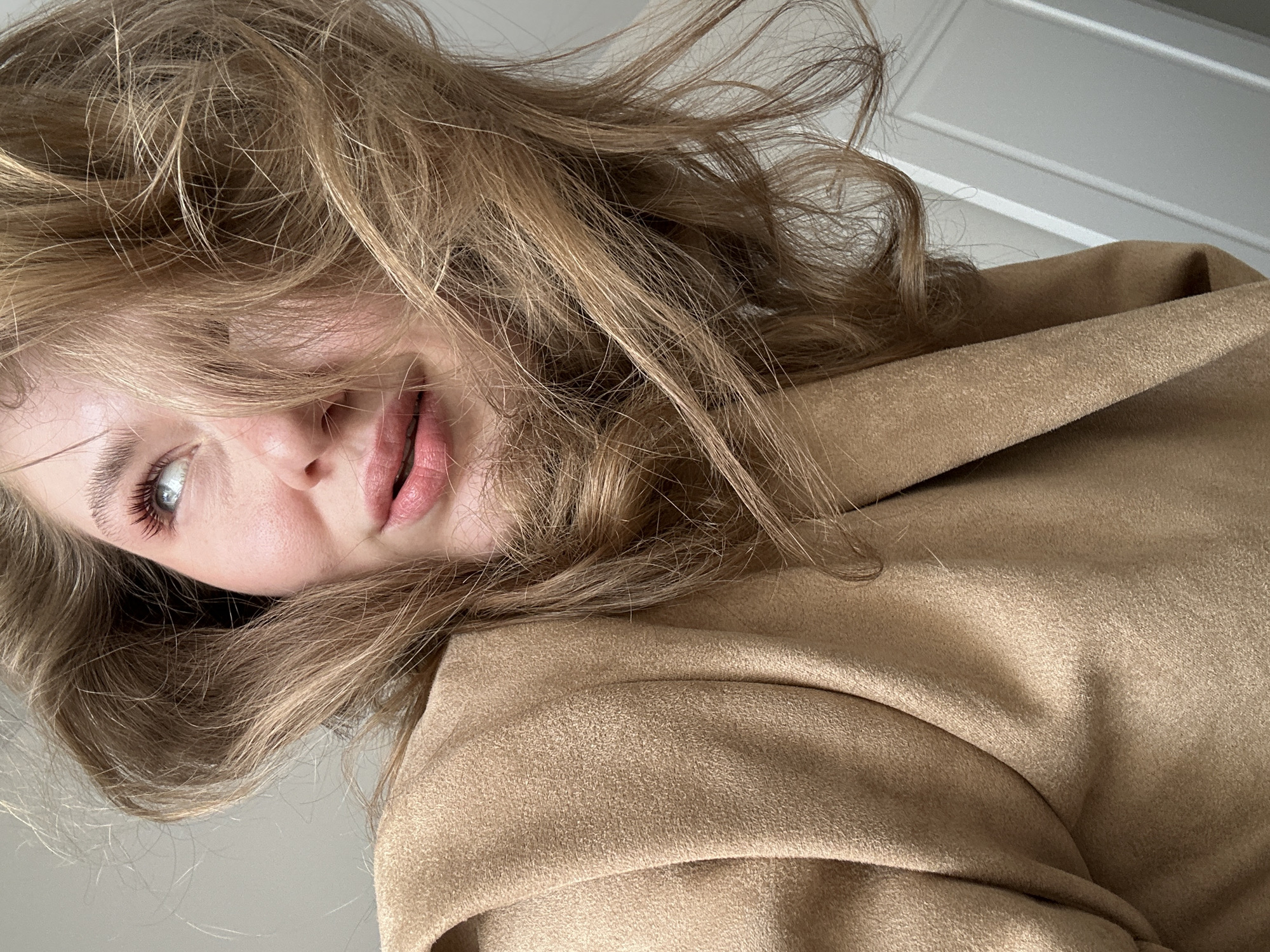 The image shows a fair-skinned person with light brown hair, blue eyes, and pink lips. The hair is tousled and partially obscures the face. The person is wearing a camel-colored coat or jacket with a prominent collar. The background is a plain, light-colored wall with a simple white doorframe visible in the upper right corner. The image is shot from a low angle, looking upwards at the person, and is slightly rotated. There is no indication of the location being Kazan, Russia in the image.