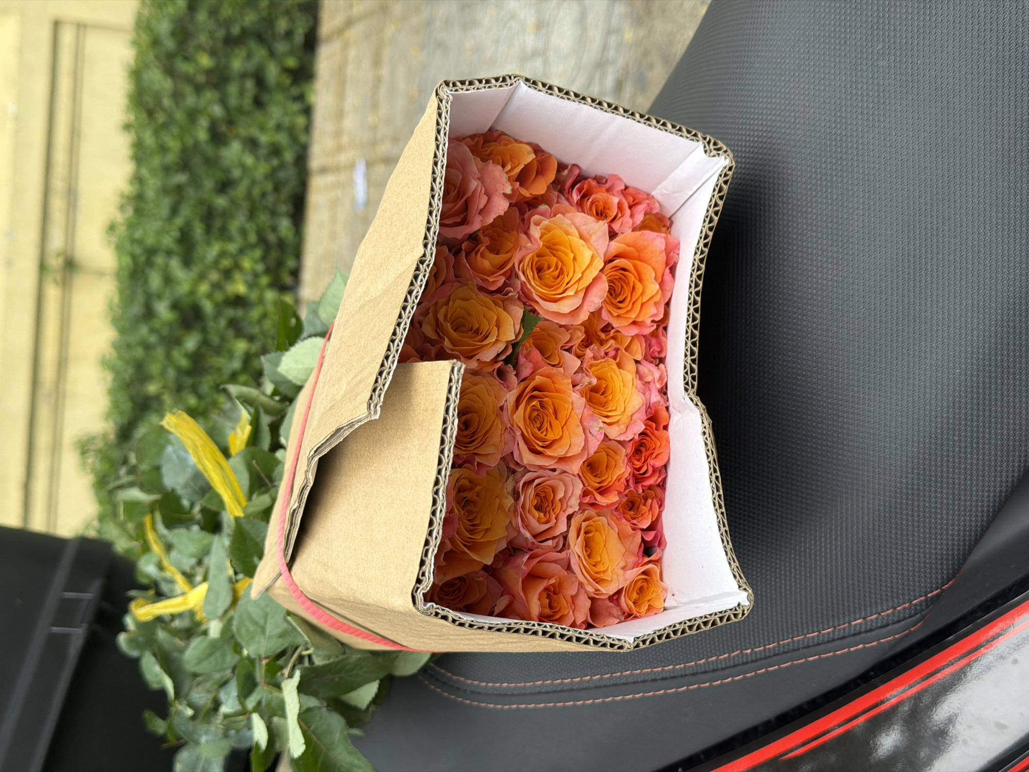 A cardboard box filled with orange and pink roses sits atop a textured black surface, likely a seat of a motorbike. The box is open, revealing rows of tightly packed flowers. Green foliage with yellow ribbons extends from the box's base. The background shows a green leafy hedge and a hint of a pale yellow wall or building. The location is likely in Nha Trang Ward, Vietnam.