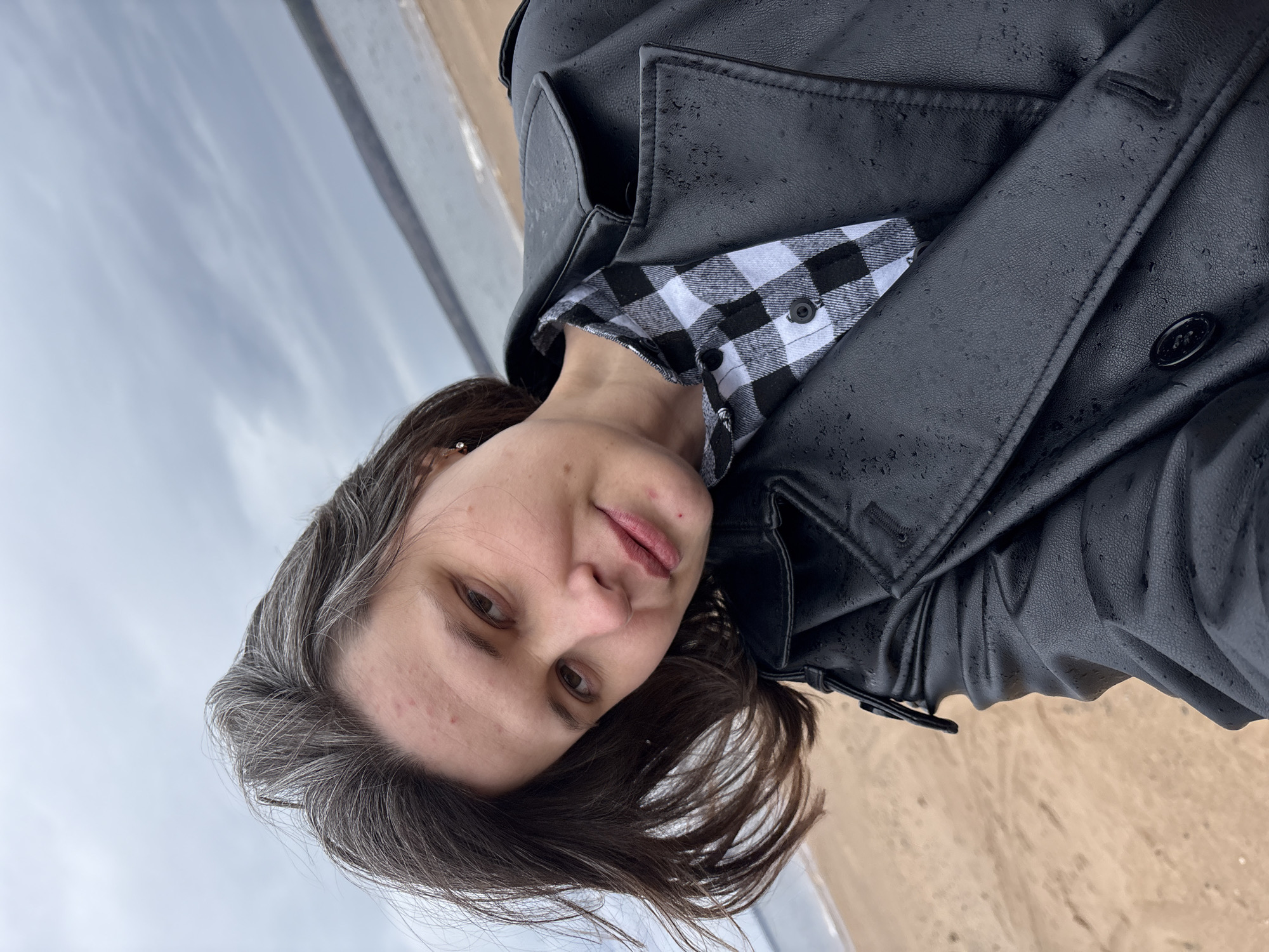 A young adult female is featured in a close-up, chest-up portrait shot outdoors. She has dark hair with noticeable grey strands and gazes directly at the viewer with a neutral to slightly serene expression. She is dressed in a black and white checkered shirt, layered with a black, leather-like coat or jacket. The surface of her coat is covered in numerous small water droplets, indicating that it is raining or has recently rained.
The background is an outdoor scene under an overcast, grey sky, visible in the upper portion of the image. The lower part of the background shows sandy or earthy ground. The scene suggests an open, natural environment, possibly a riverbank or beach, consistent with the location of Chaikovsky, Russia, which is situated on the Kama River. There is no visible text in the image. The overall impression is one of a quiet moment captured during damp, cool weather.