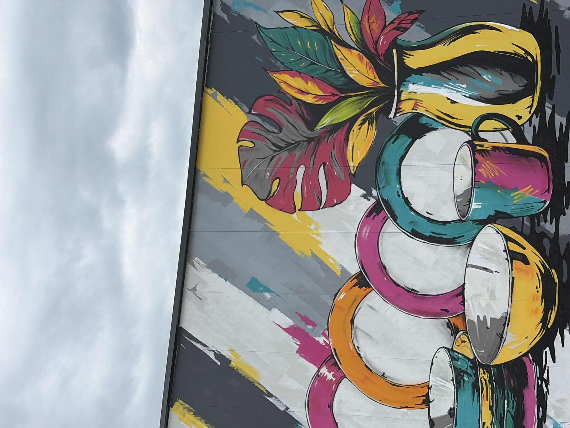 The image depicts a mural on the side of a building, likely in Sofia, Bulgaria, though this cannot be confirmed from the image alone. The mural is a still life of a vase with leaves and a stack of plates and a mug. The vase is a yellow and black, organic shaped vase with various green, red, yellow, and orange leaves protruding from the top. There is also a large red and gray monstera leaf. Below the vase is a magenta and blue mug, followed by a stack of plates. The plates are painted with blue, magenta, yellow and orange rings. The background of the mural is gray with yellow, white, blue, magenta, and gray diagonal strokes. The building wall is gray with visible seams. The upper left of the frame shows an overcast sky.